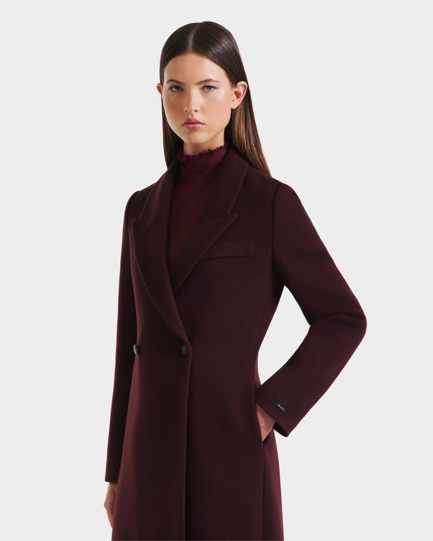 A woman with straight brown hair wears the Rudsak LYNWOOD - 8125539 Bordeaux, a dark burgundy women’s long tailored double-breasted coat, over a matching high-neck top, standing against a plain white background.