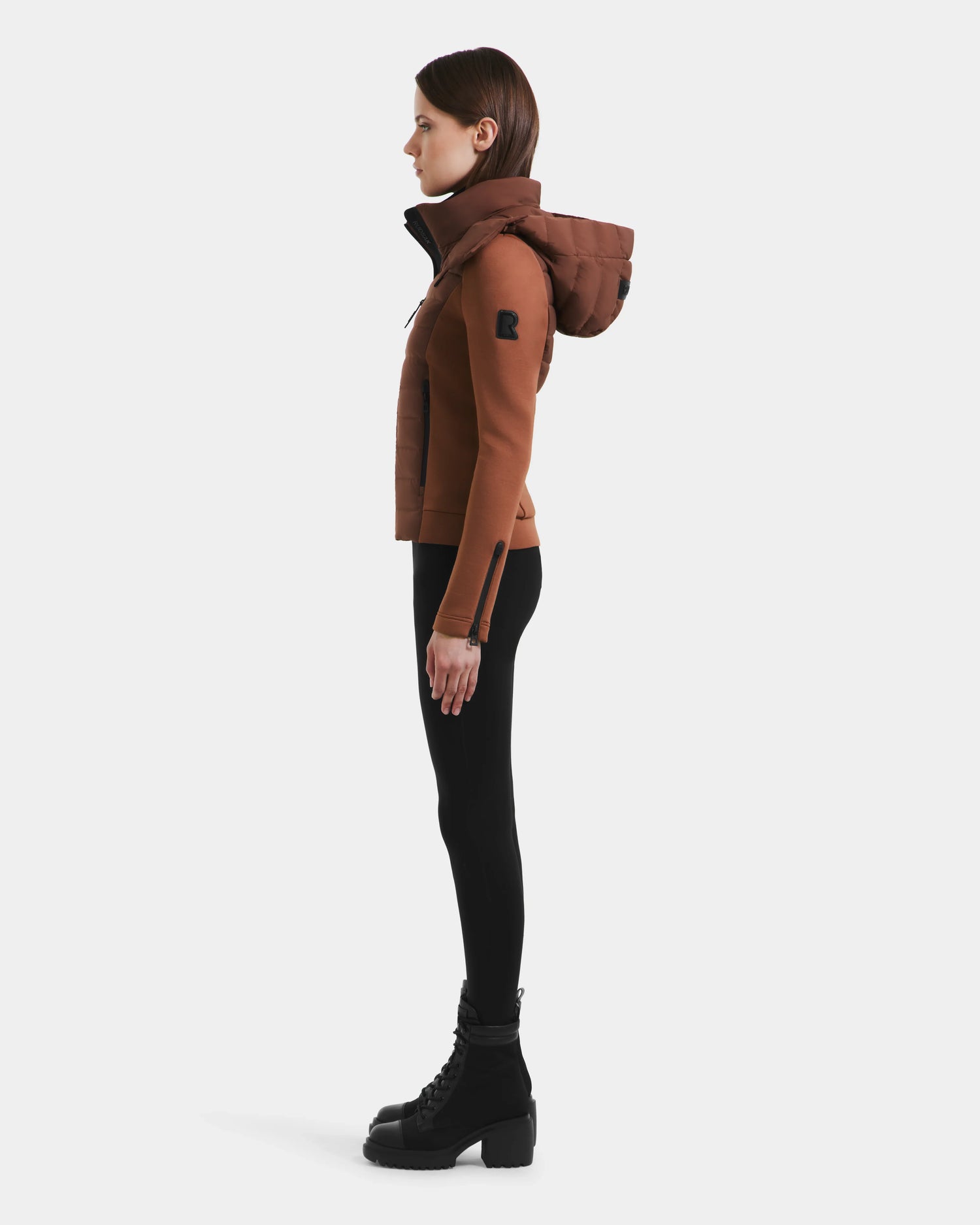 A woman stands in profile wearing the Rudsak MABS - 8125055 Rust lightweight activewear jacket with black leggings and lace-up boots against a plain white background.