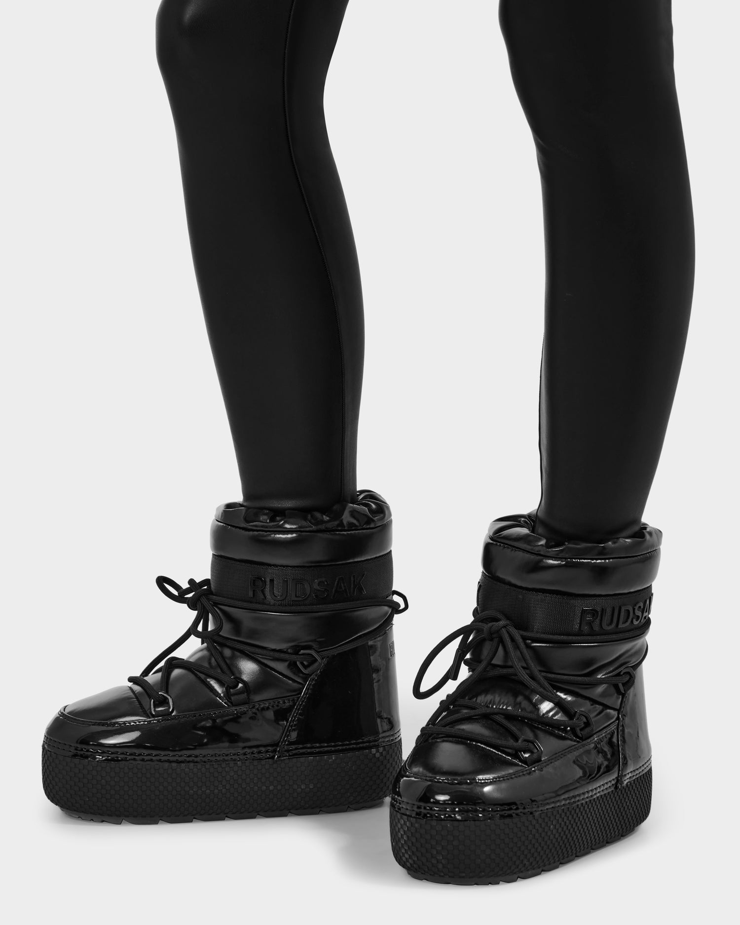A person wears MACE - 8223500 Black boots by Rudsak, featuring a glossy water-repellent finish, thick soles, black laces, faux fur lining for warmth, front RUDSAK branding, and a trendy short moon boot silhouette with fitted leggings.