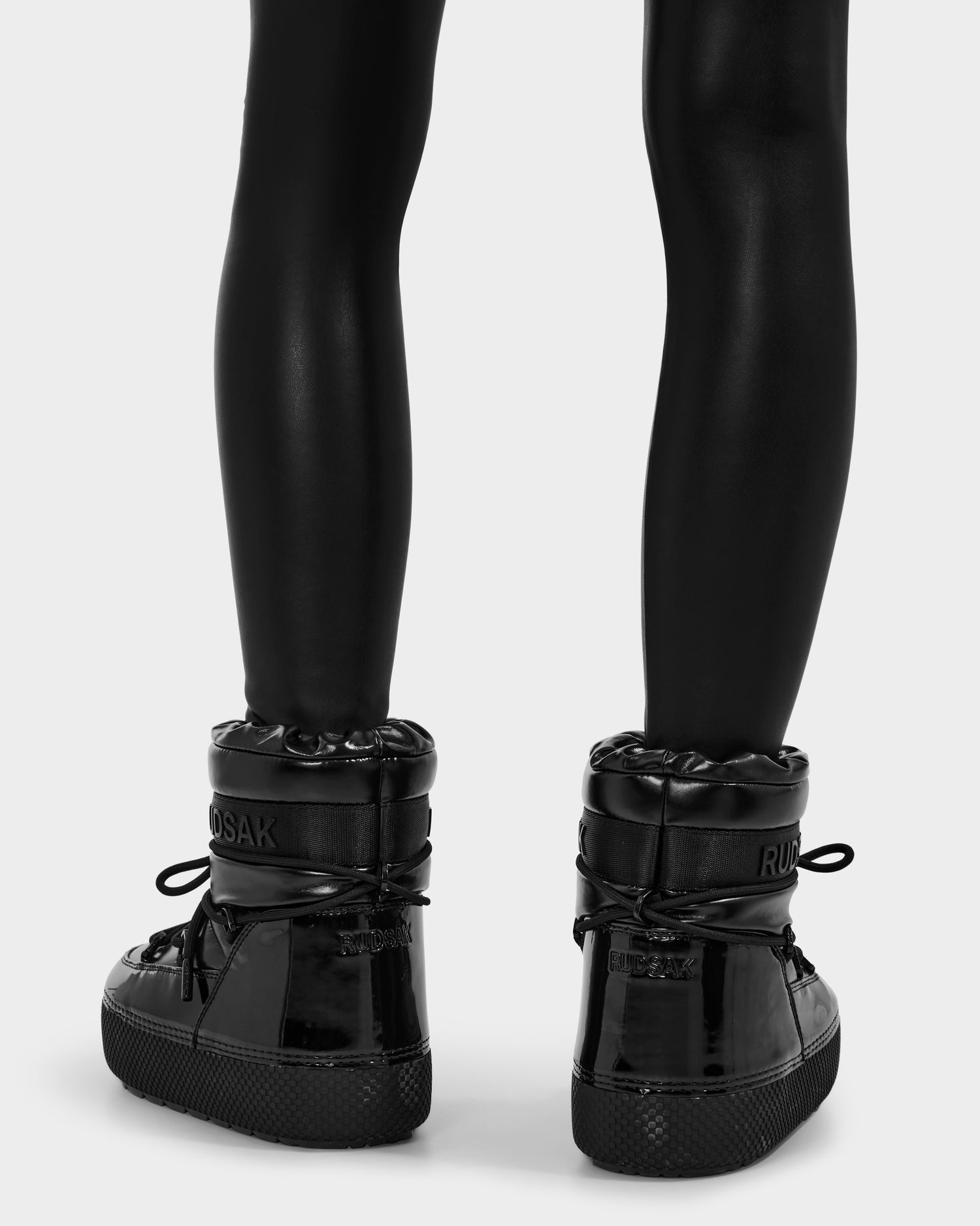 A person wearing shiny black leggings and Rudsak MACE - 8223500 Black glossy water-repellent women’s short moon boots with thick soles, viewed from behind against a plain light background.