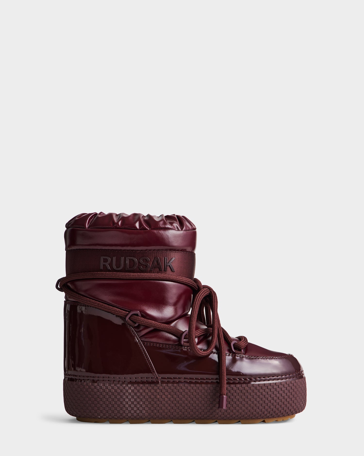 The Rudsak MACE - 8223500 Bordeaux is a maroon high-top winter boot with a shiny patent finish, thick sole, rounded toe, front laces, quilted insulated look, faux fur lining for warmth, and RUDSAK branding on the upper shaft.