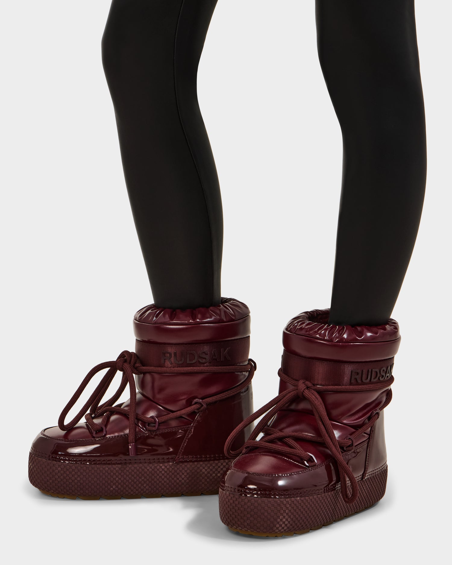 A person wearing Rudsak's MACE - 8223500 Bordeaux boots with thick soles, faux fur lining, and large laces, paired with black leggings. The photo highlights the stylish water-repellent winter footwear on their lower legs.