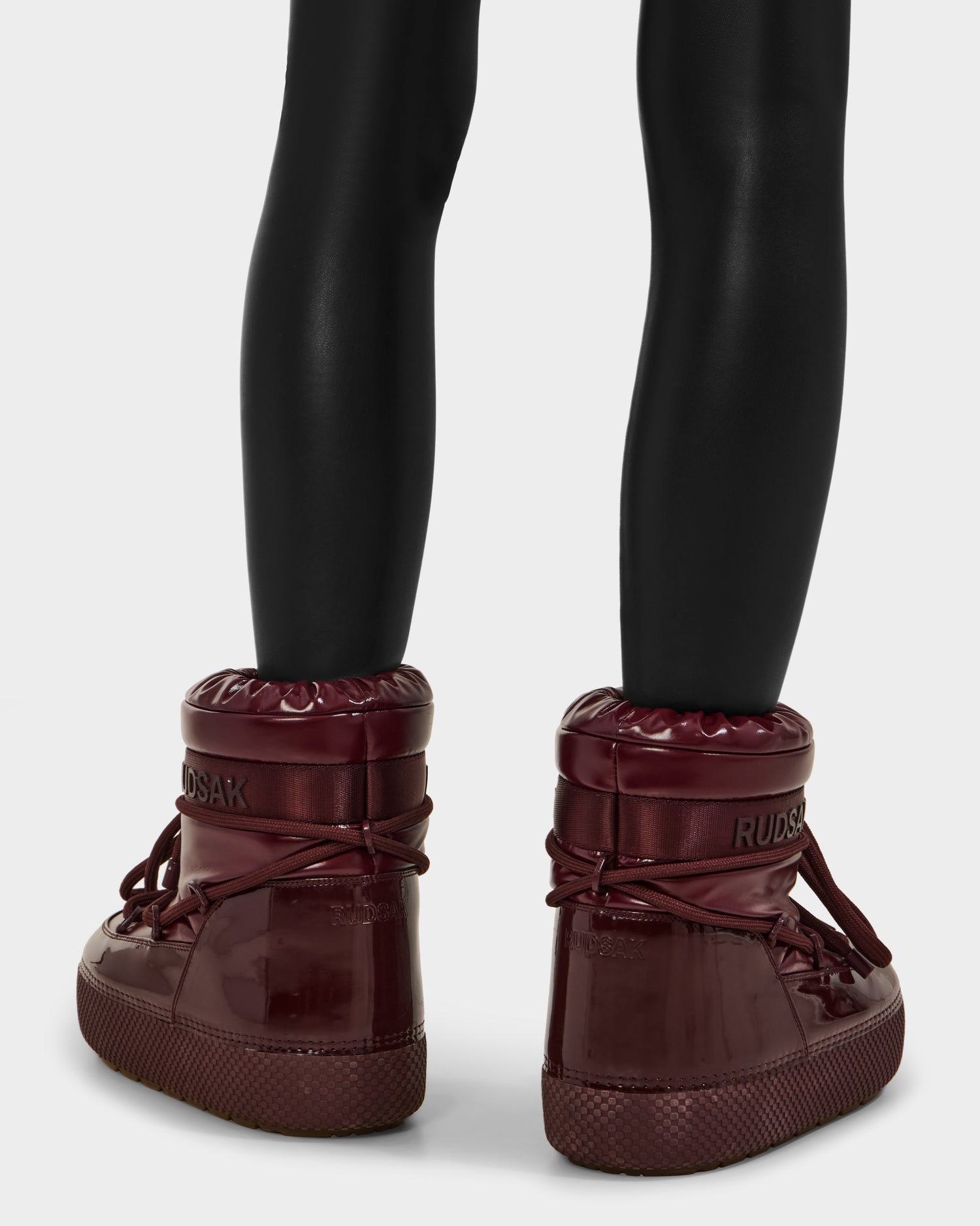 Someone is shown from behind against a plain background, wearing black leggings and shiny, dark red Rudsak MACE - 8223500 Bordeaux high-top boots with thick soles, laces, and a water-repellent finish.