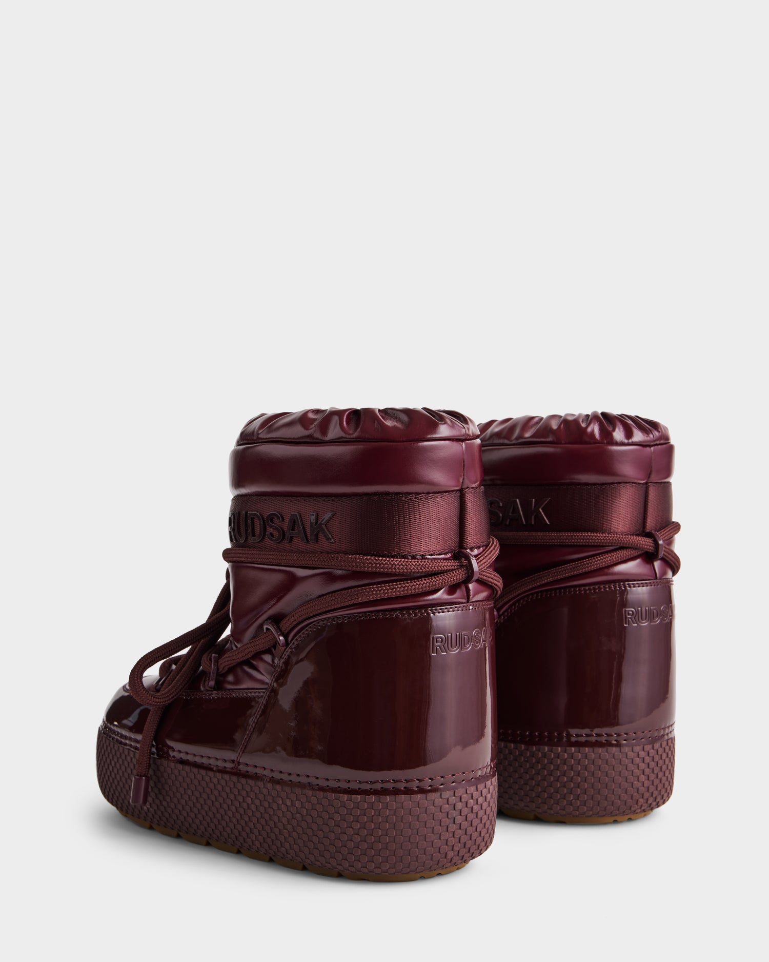 The Rudsak MACE - 8223500 Bordeaux women's short moon boots feature a shiny maroon finish, thick soles, rounded toes, drawstring closures, and cozy faux fur lining, displayed on a plain light gray background.