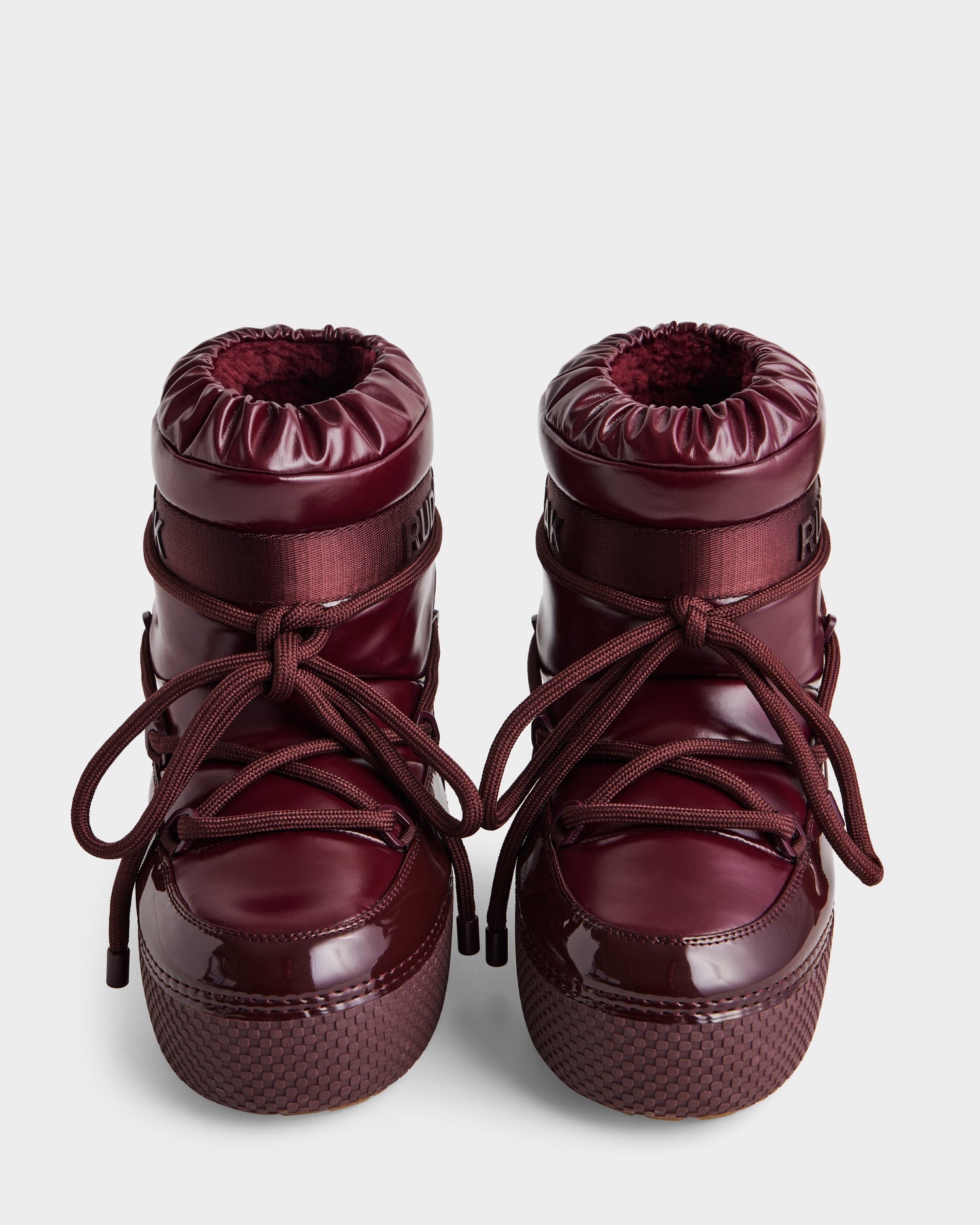 The Rudsak MACE - 8223500 Bordeaux women's short moon boots feature shiny burgundy puffy uppers, thick soles, rounded toes, and wrap-around laces. The boots stand upright on a plain, light background.