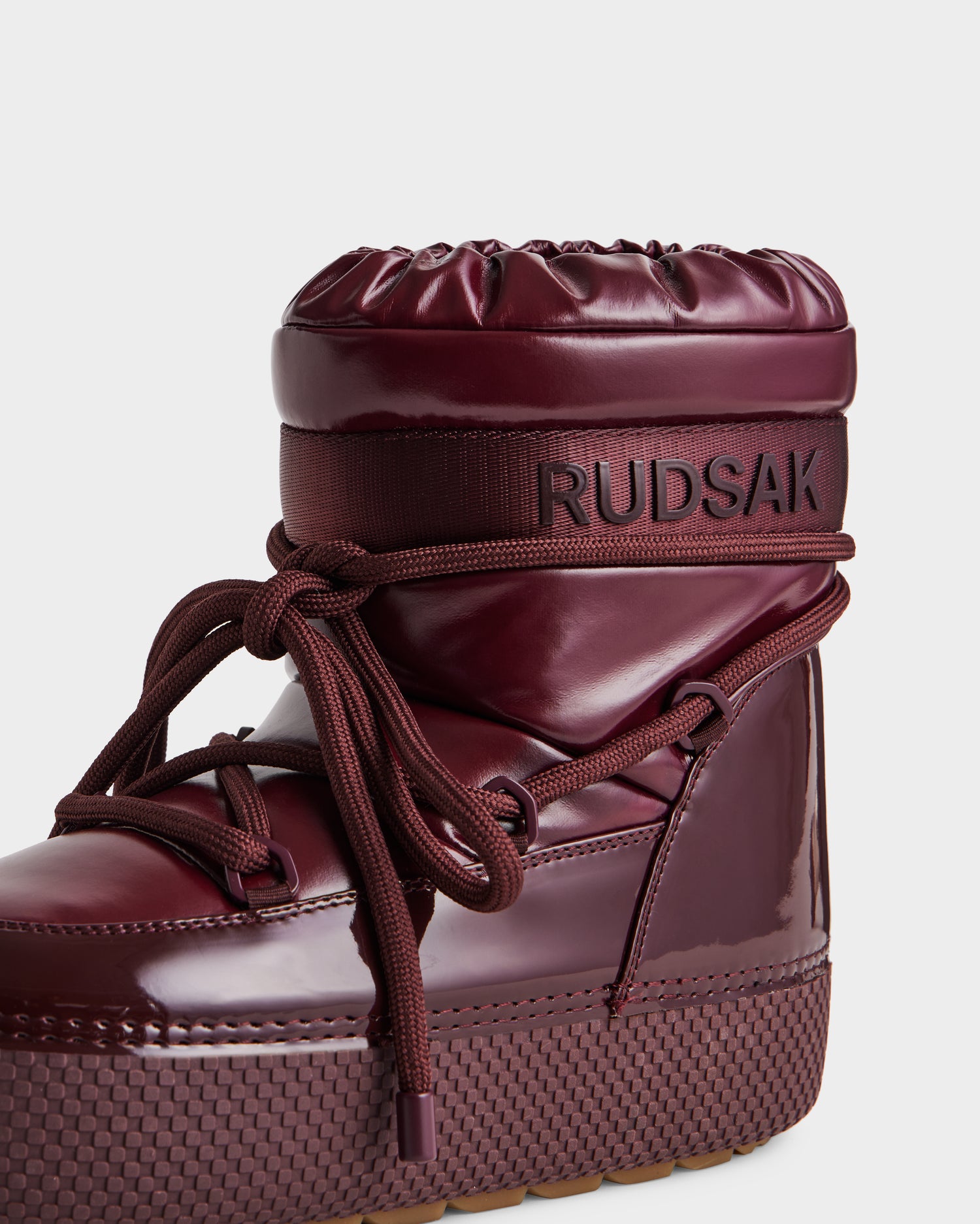 Close-up of the Rudsak MACE - 8223500 Bordeaux women's short moon boot, featuring glossy water-repellent material, textured sole, thick laces, padded faux fur-lined upper, and Rudsak branding on a horizontal strap.