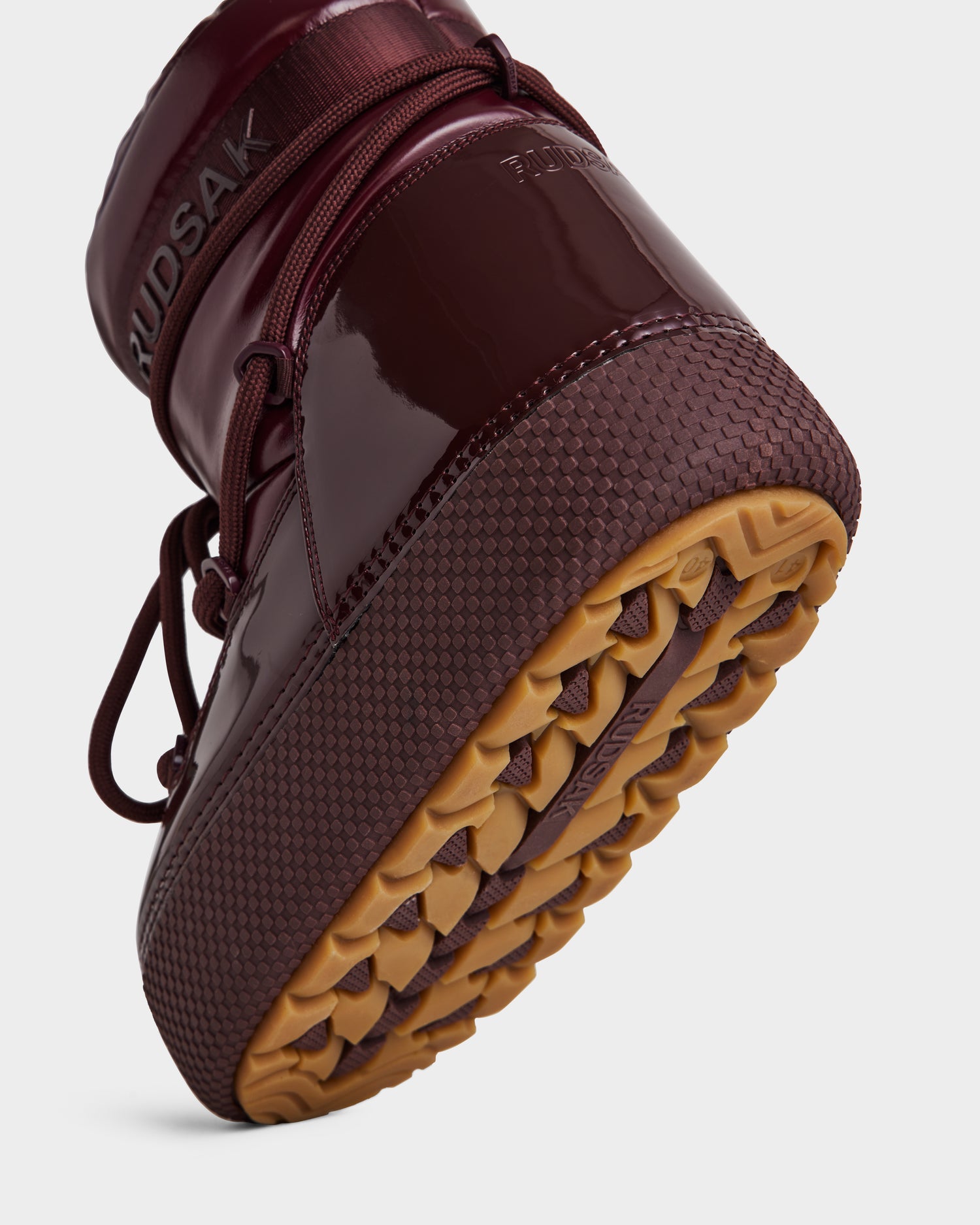 Close-up of the Rudsak MACE - 8223500 Bordeaux women's boot with glossy maroon finish, tan textured tread, laces, and embossed branding on the heel. Features faux fur lining for warmth. Set against a plain white background.