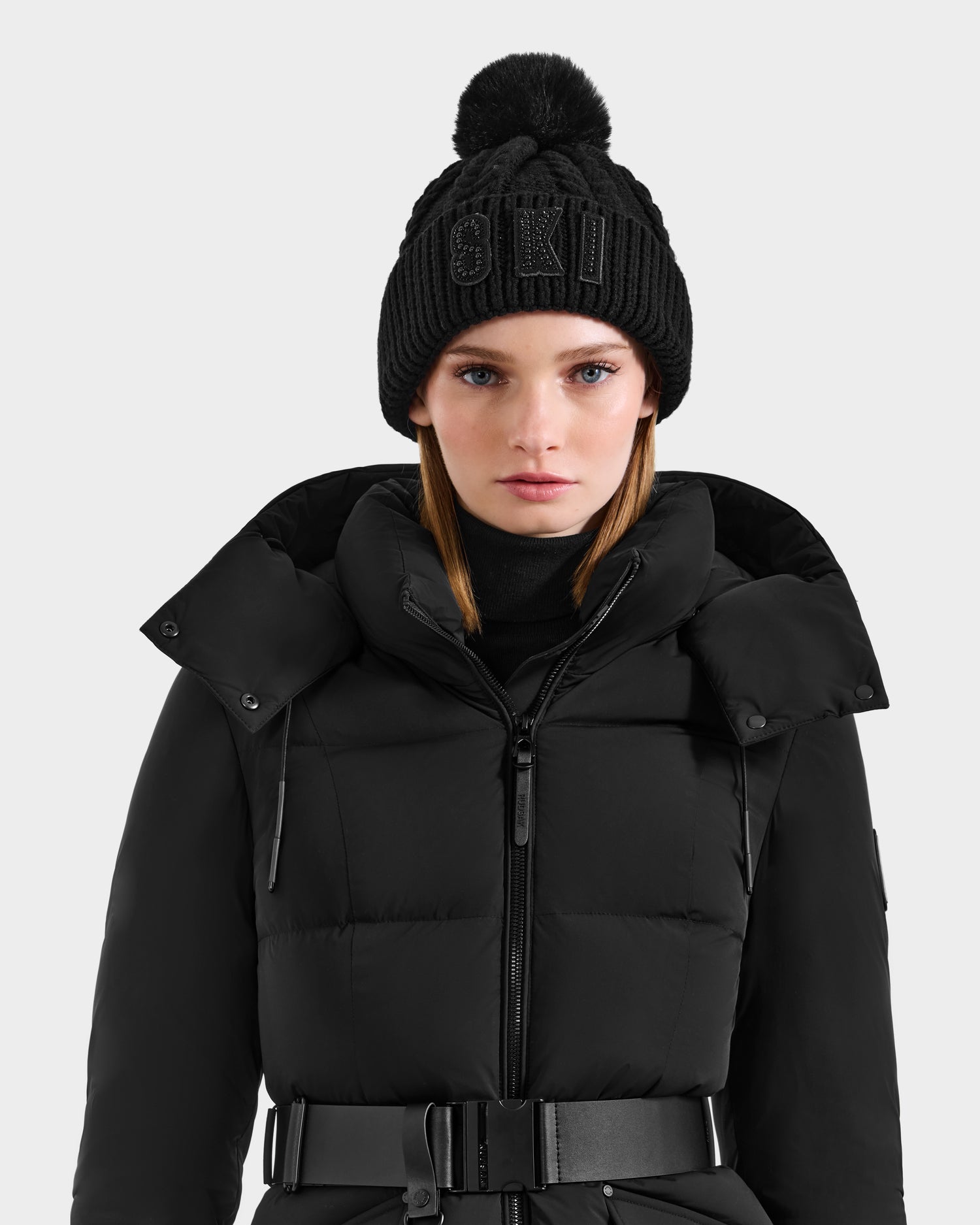 A woman in the Rudsak MAE - 8425550 Black puffer jacket, featuring a high collar and belted waist, wears a cable knit tuque with a crystal SKI logo and faux fur pom pom, standing against a plain light background.