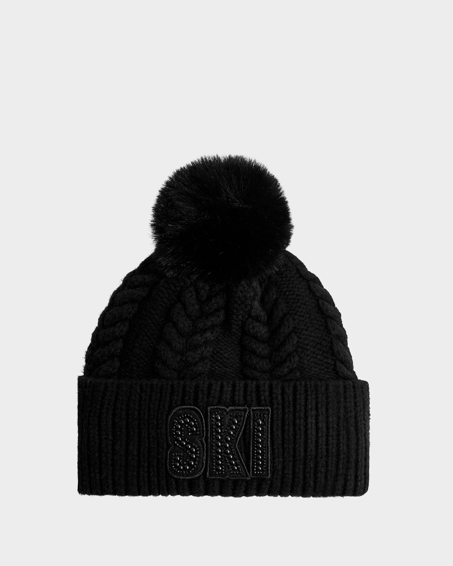 The Rudsak MAE - 8425550 Black is a black cable knit tuque with a large faux fur pom pom and SKI in bold capitals on the folded brim.