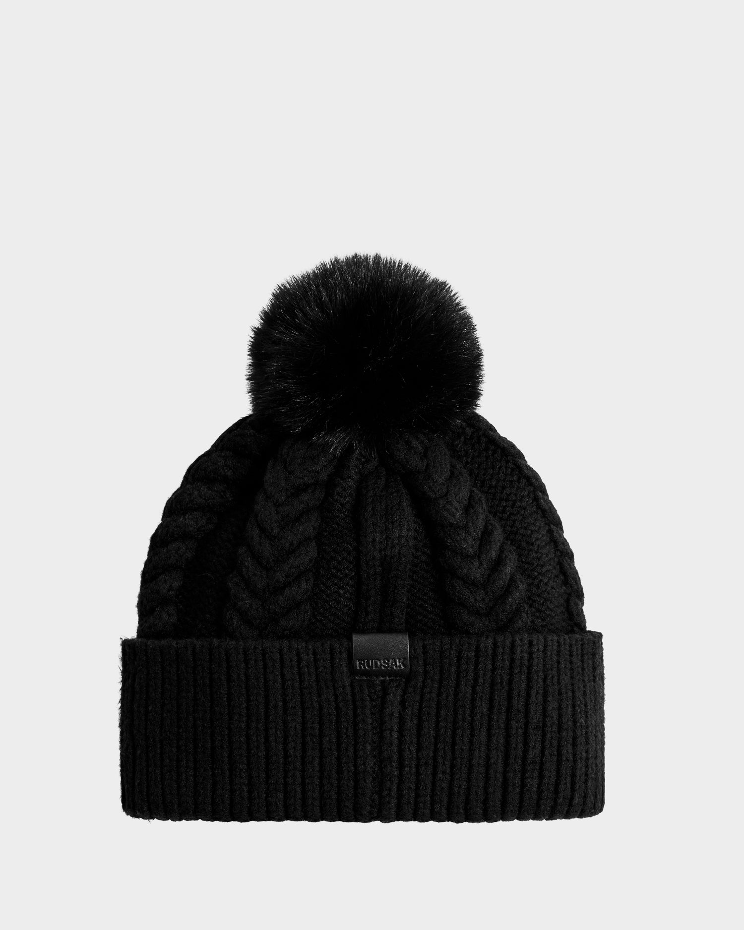 The Rudsak MAE - 8425550 Black is a black cable knit tuque featuring a folded brim, chunky pattern, and a large black faux fur pom pom on top.