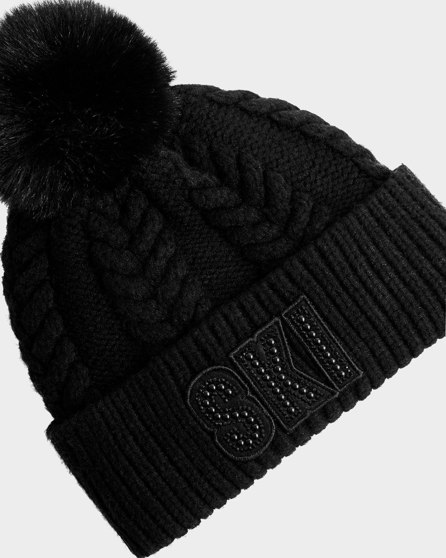 The Rudsak MAE - 8425550 Black is a black cable knit tuque with a large faux fur pom pom and 'SKI' in bold, textured letters with decorative studs on the front folded brim.
