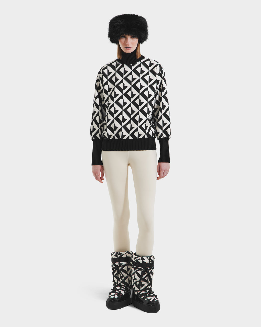 A person wearing Rudsaks MAGGIE - 8525645 Vanilla Monogram boots stands against a plain white background, paired with an alpine knit sweater, cream leggings, and a black fur hat.