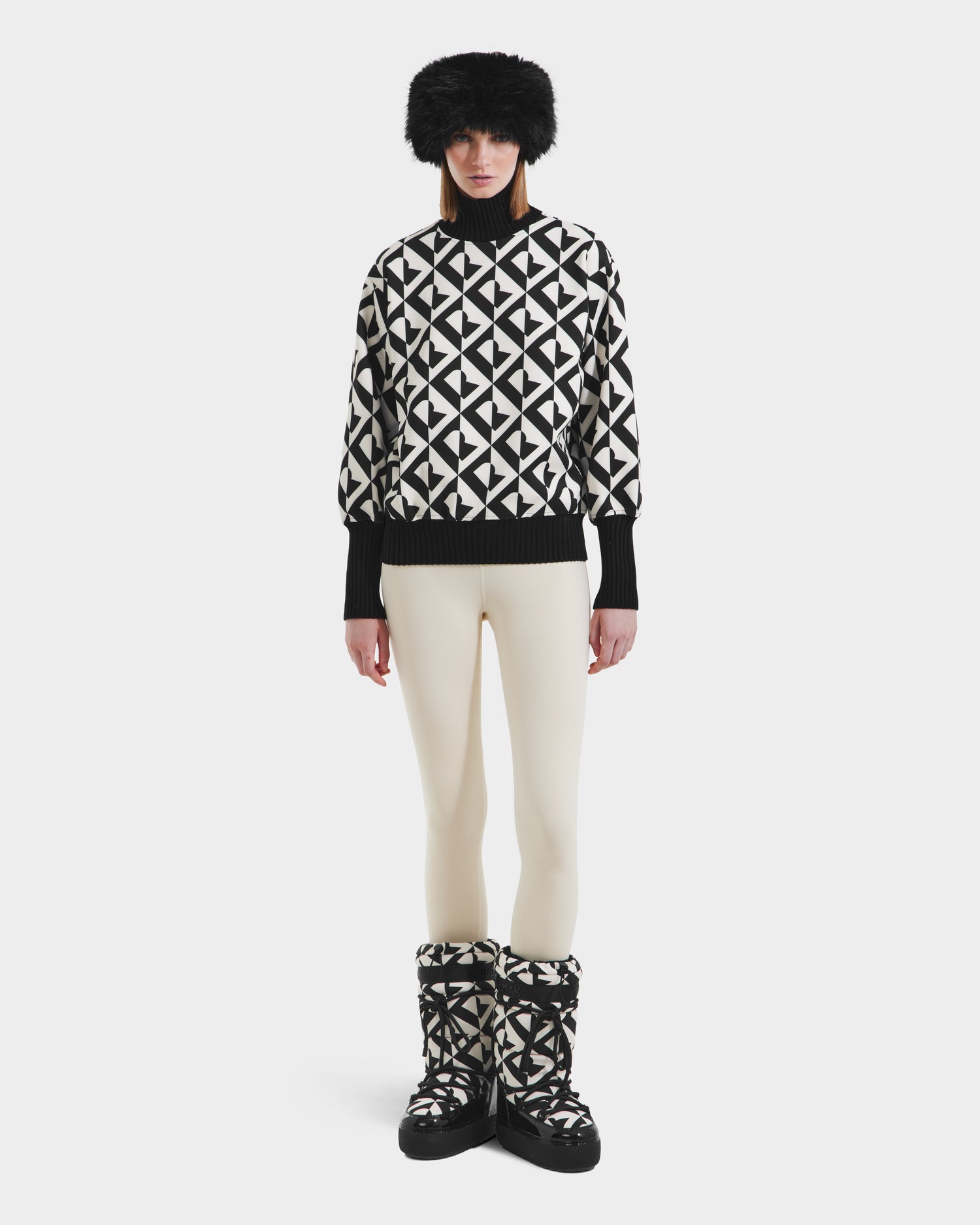 A person wearing Rudsaks MAGGIE - 8525645 Vanilla Monogram boots stands against a plain white background, paired with an alpine knit sweater, cream leggings, and a black fur hat.