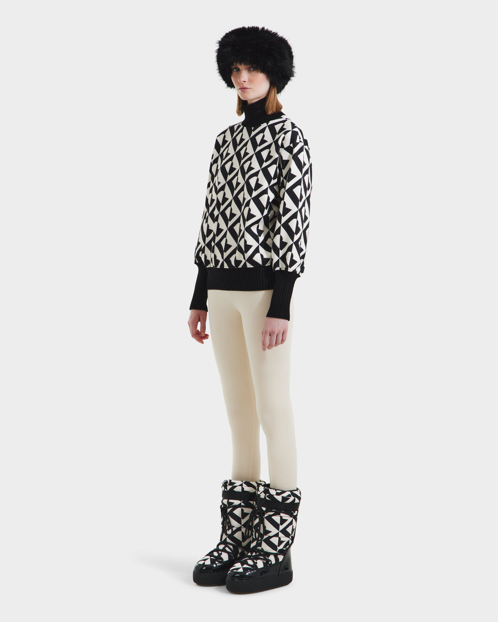 A person stands slightly to the side, wearing Rudsaks MAGGIE - 8525645 Vanilla Monogram winter boots, paired with a black fur hat, an alpine knit womens ski sweater with black-and-white patterns, and cream leggings against a white background.