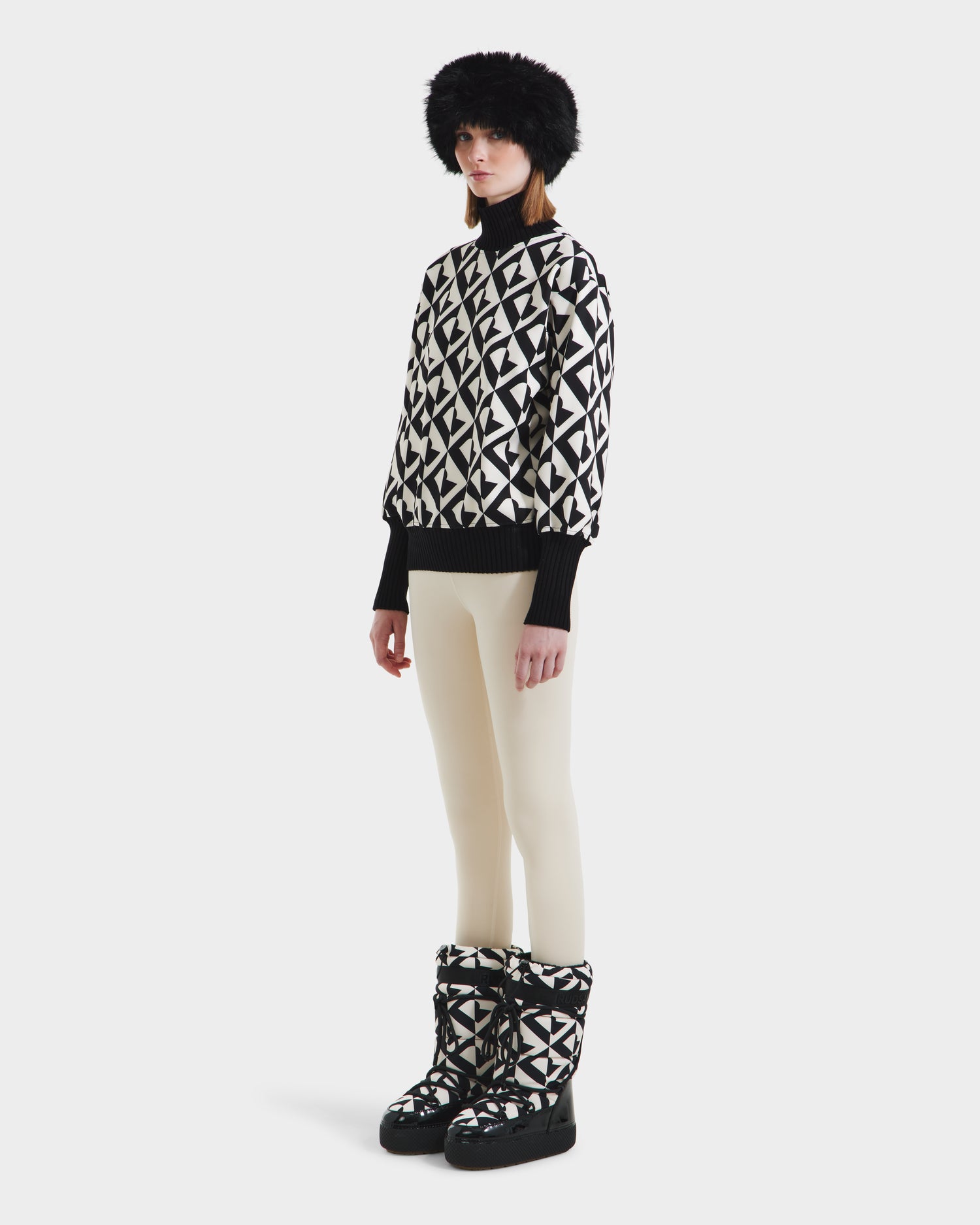 A person stands slightly to the side, wearing Rudsaks MAGGIE - 8525645 Vanilla Monogram winter boots, paired with a black fur hat, an alpine knit womens ski sweater with black-and-white patterns, and cream leggings against a white background.