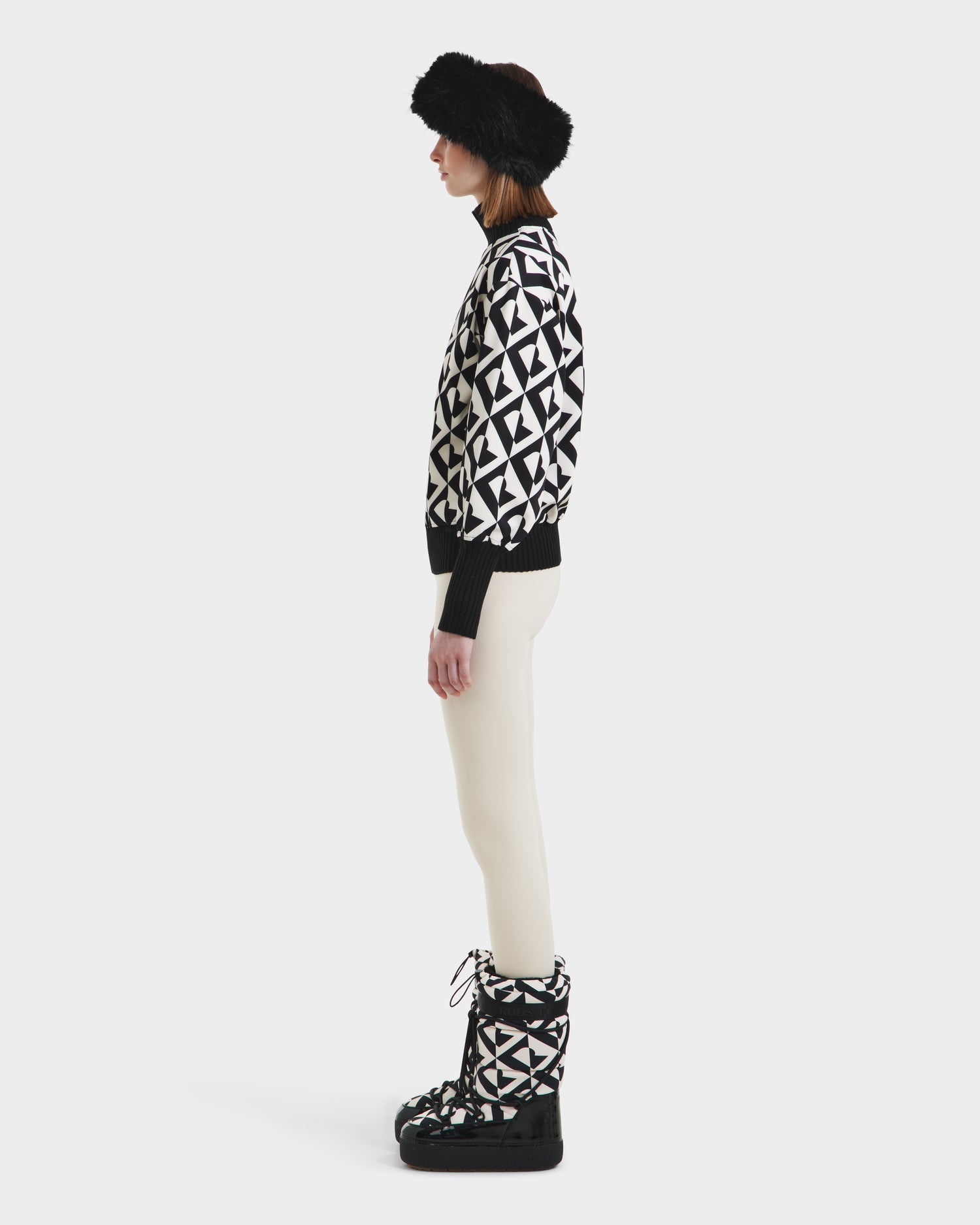 A person stands in profile against a plain white background, wearing Rudsaks MAGGIE - 8525645 Vanilla Monogram boots, paired with a black fur hat, a black and white geometric cotton ski sweater, and white pants.