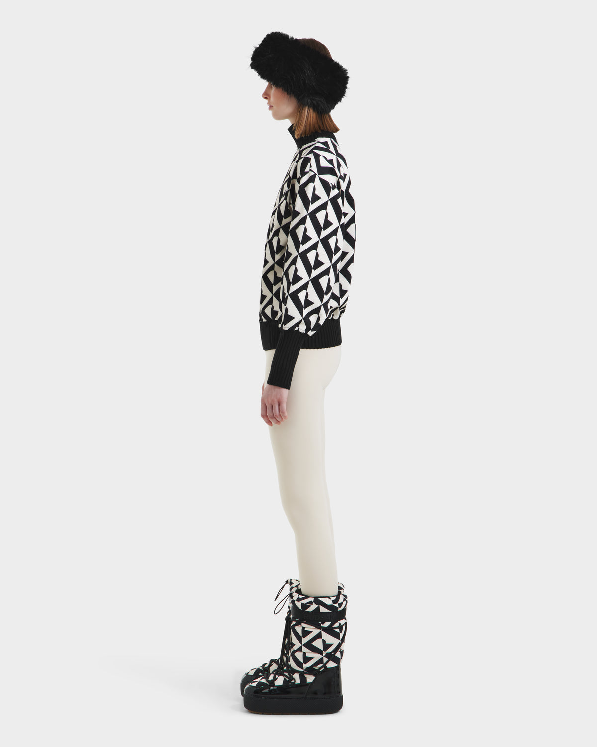 A person stands in profile against a plain white background, wearing Rudsaks MAGGIE - 8525645 Vanilla Monogram boots, paired with a black fur hat, a black and white geometric cotton ski sweater, and white pants.