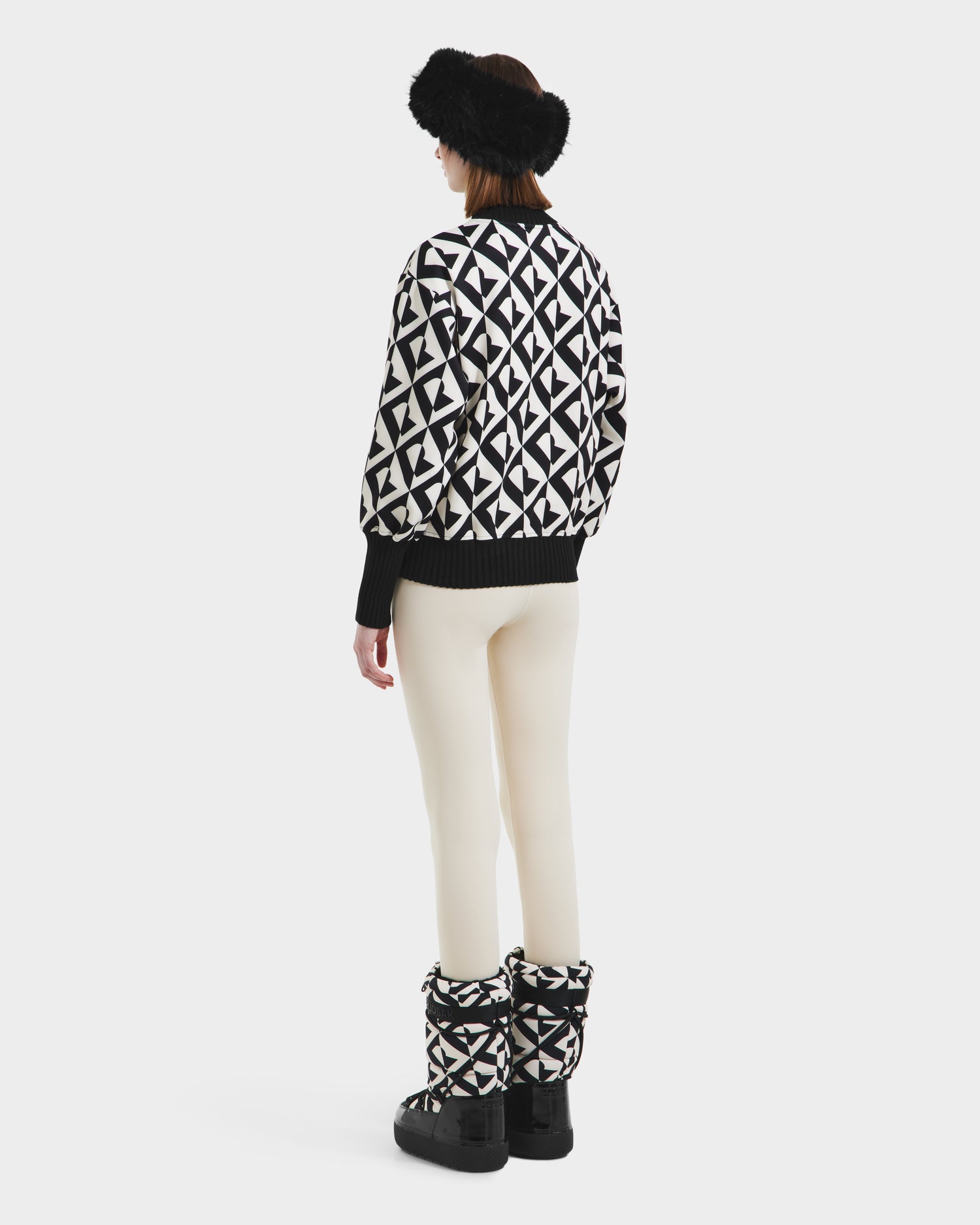 A woman faces away against a white background, wearing Rudsaks MAGGIE - 8525645 Vanilla Monogram cream pants, paired with a black and white alpine knit sweater, matching boots, and a black fur headband.