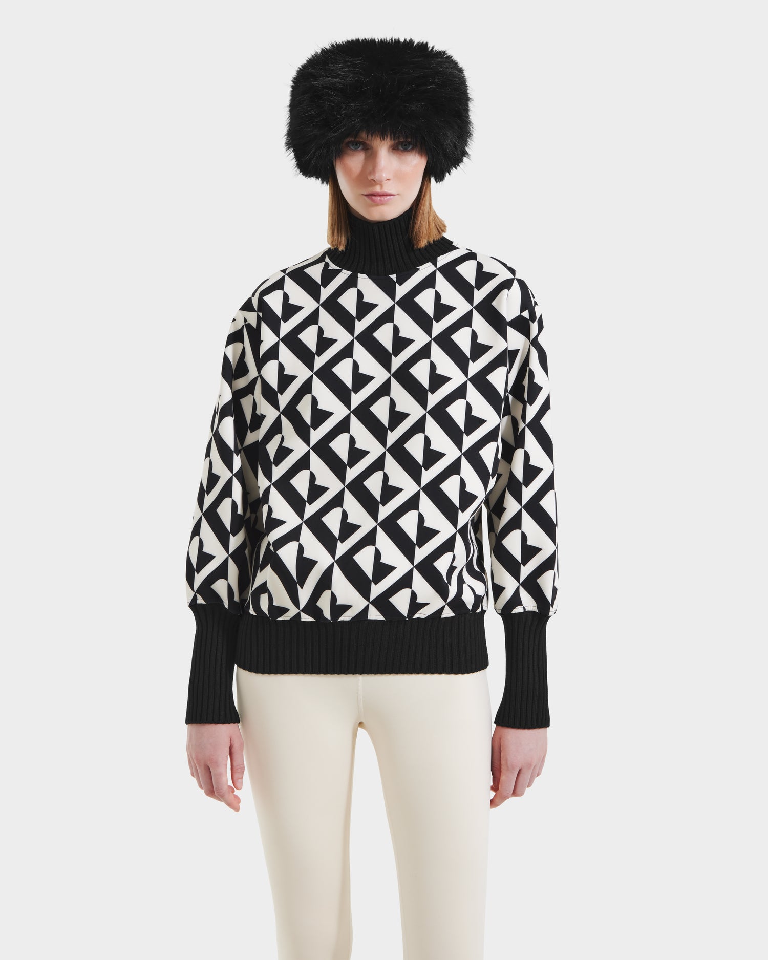 A woman wears the Rudsak MAGGIE - 8525645 Vanilla Monogram women’s ski sweater with black cuffs and collar, a black fur hat, and off-white pants, standing against a plain white background.