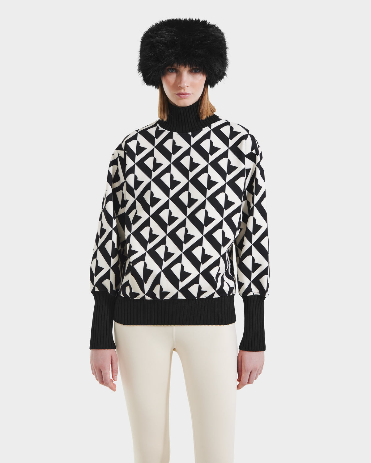 A woman wears the Rudsak MAGGIE - 8525645 Vanilla Monogram women’s ski sweater with black cuffs and collar, a black fur hat, and off-white pants, standing against a plain white background.