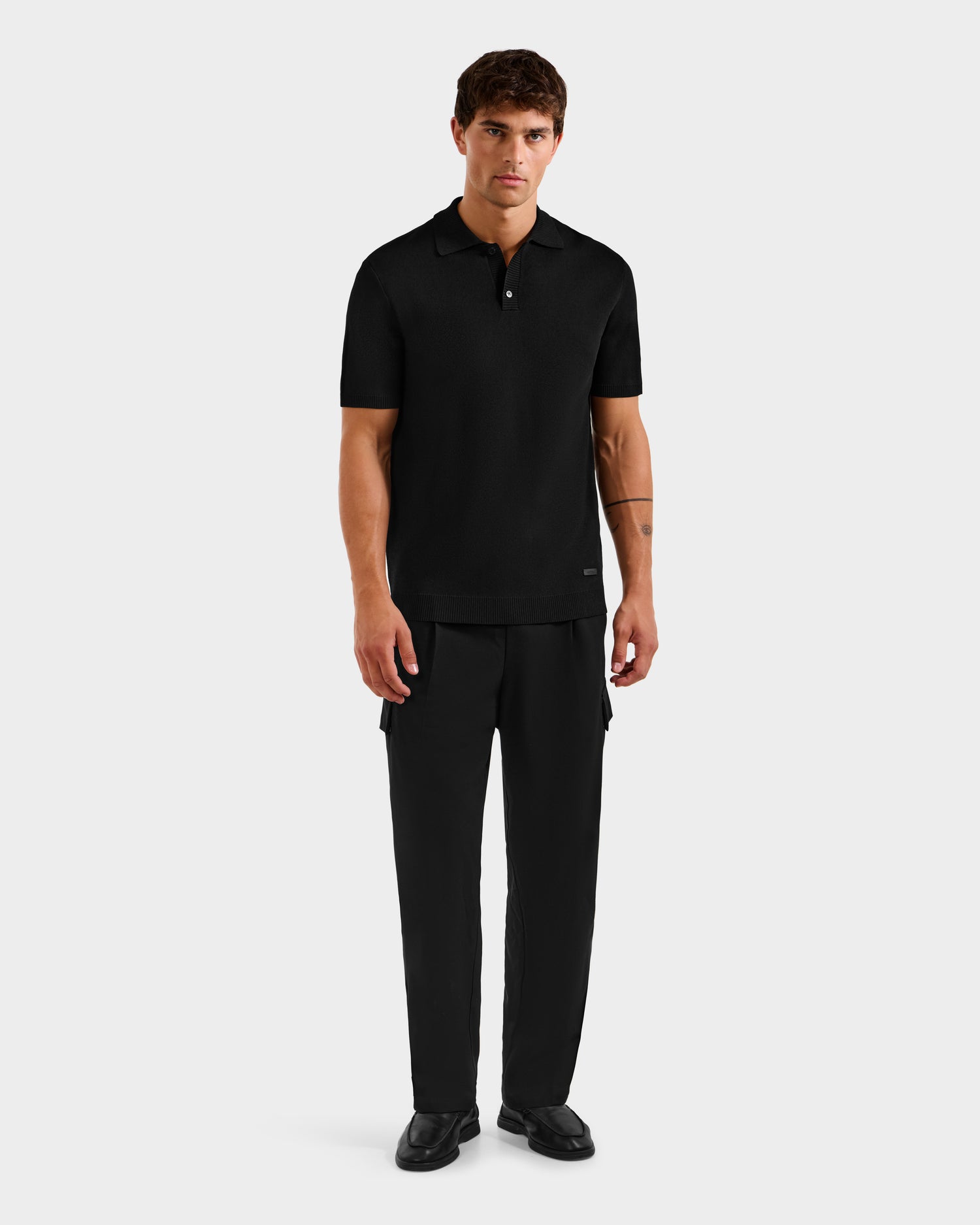 A man stands facing forward, wearing the Rudsak MAISEN - 6524518 Black polo with matching black trousers and shoes, set against a plain light gray background.