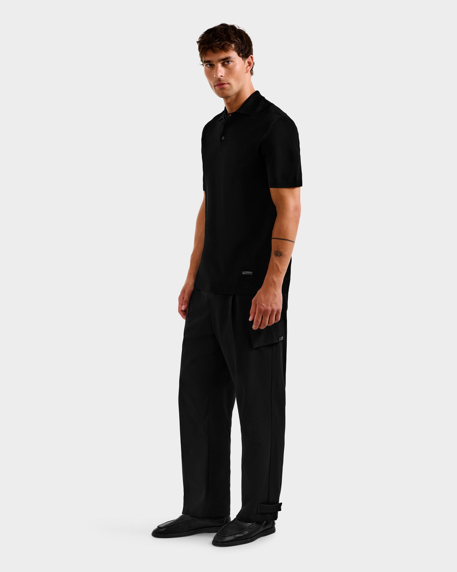 A man with short brown hair and a thin tattoo on his arm stands against a plain light background, wearing Rudsak’s MAISEN - 6524518 Black polo and loose black pants, paired with black shoes—a modern men’s wardrobe classic.