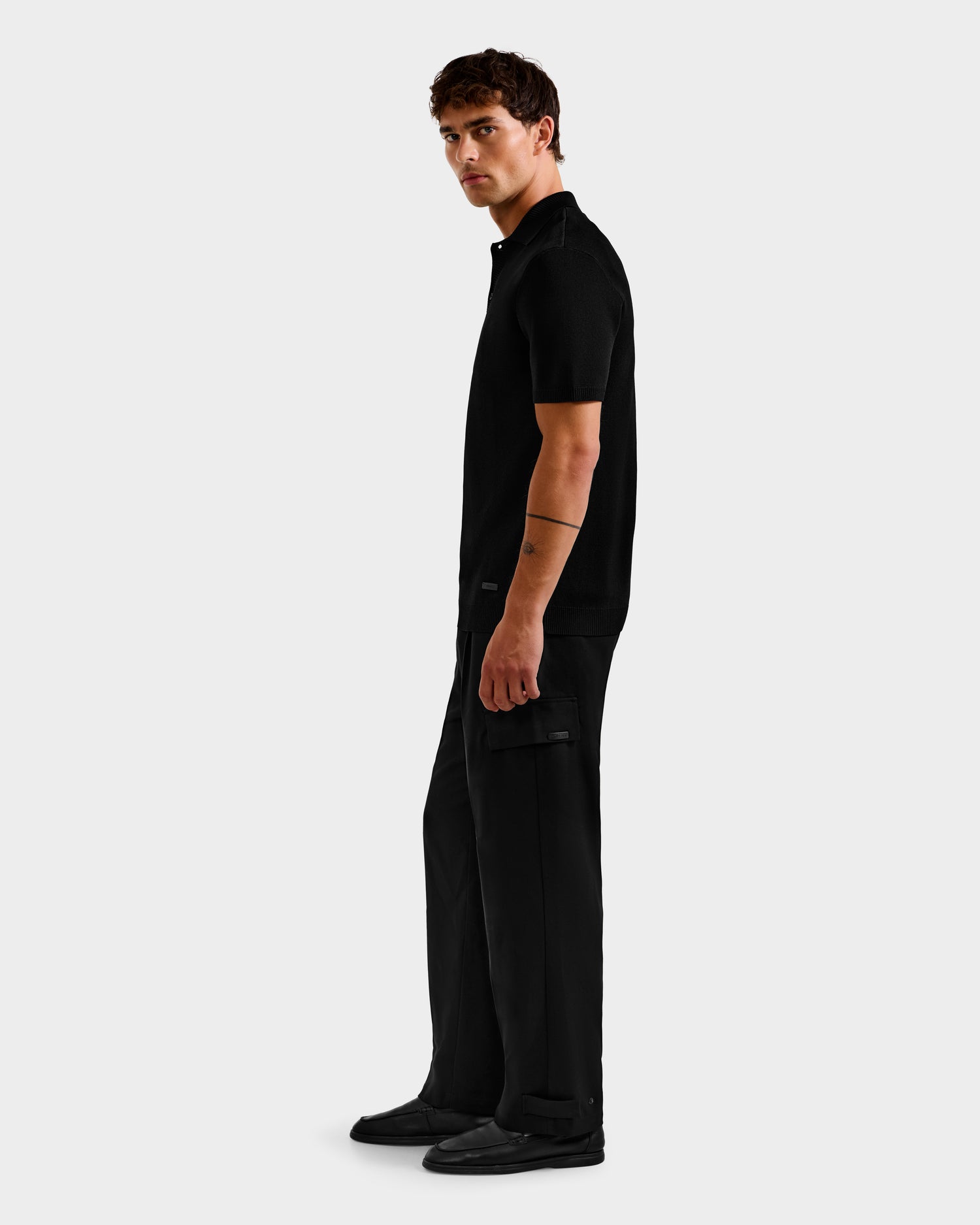 A man faces sideways wearing the MAISEN - 6524518 Black polo and cargo pants by Rudsak, paired with matching shoes—an effortlessly stylish outfit for the modern man—set against a solid light gray background.