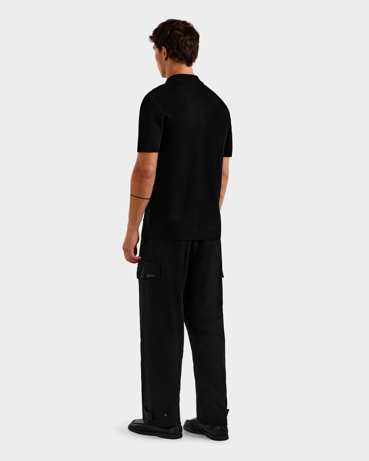 A person stands facing away from the camera, wearing the Rudsak MAISEN - 6524518 Black short-sleeve polo with black cargo pants and shoes against a plain light gray background—a sleek look for the modern men's wardrobe.