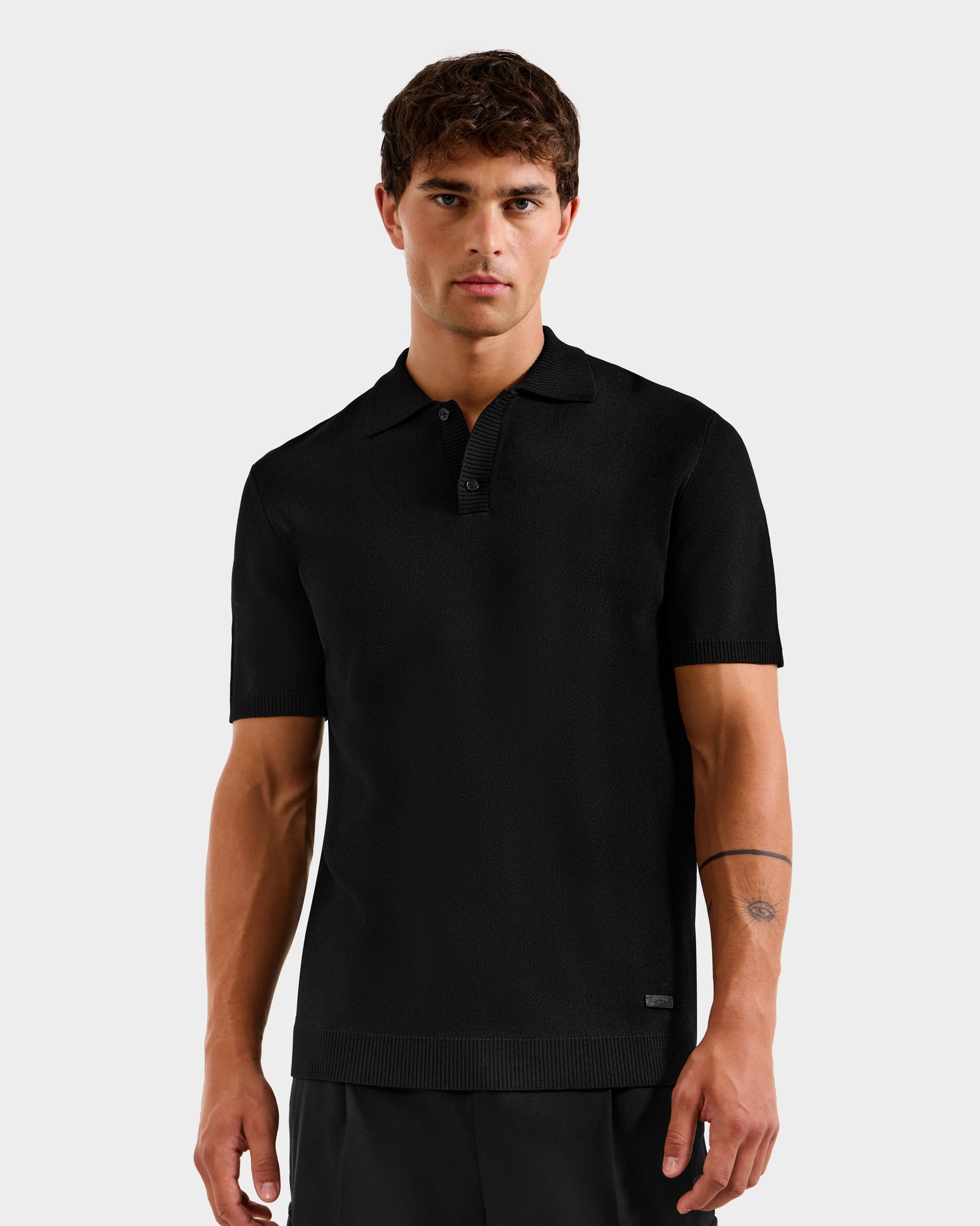 A young man with short brown hair wears the Rudsak MAISEN - 6524518 Black polo and black pants, standing against a plain white background with a neutral expression. A simple line tattoo is visible on his left forearm.