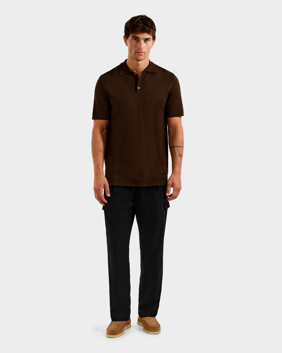 A man stands facing forward against a plain light gray background, wearing Rudsak's MAISEN - 6524518 Mocha short-sleeve men's knit polo, paired with black pants and brown shoes.