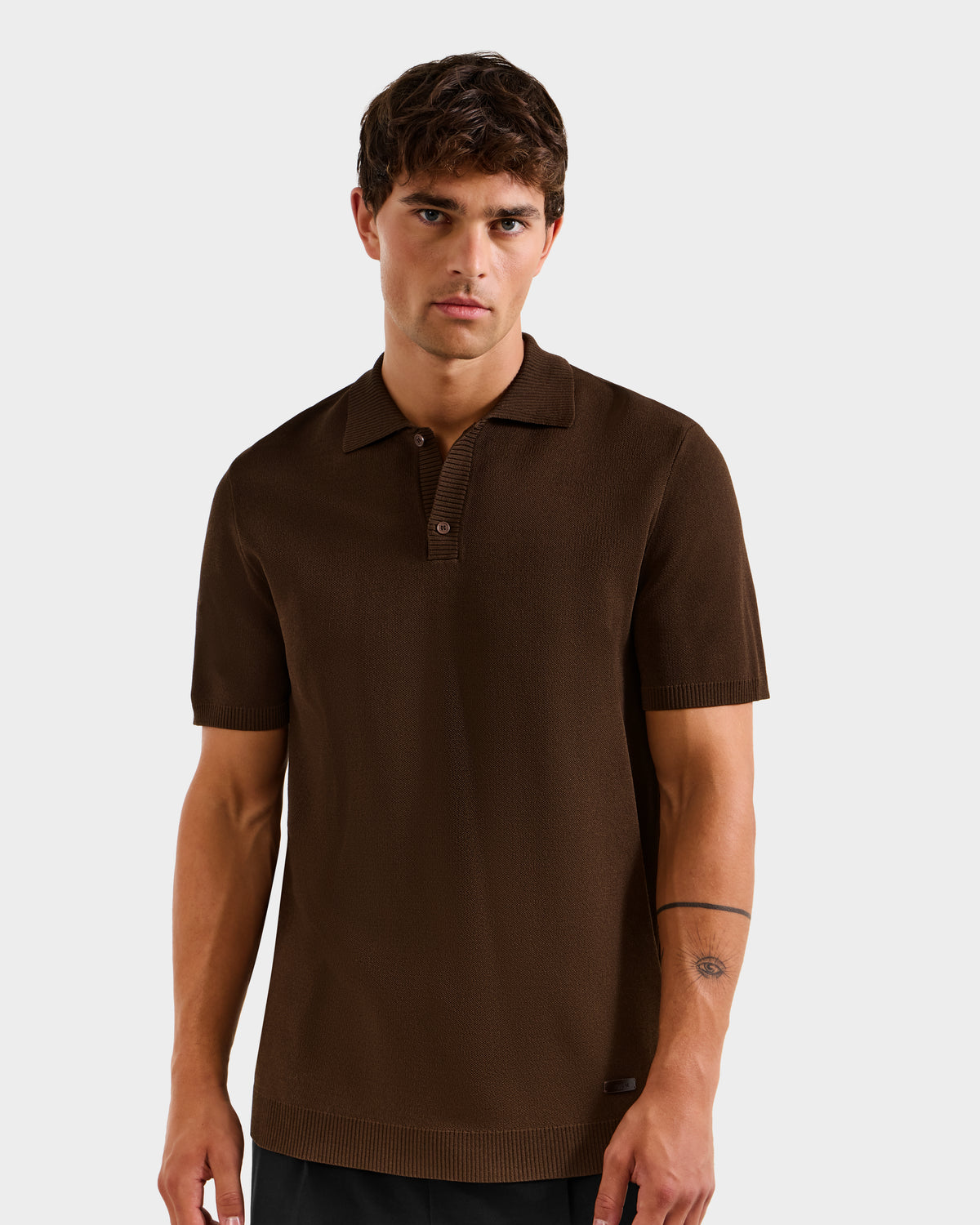 A man with short brown hair wears the Rudsak MAISEN - 6524518 Mocha men's knit polo and dark pants, standing against a plain light gray background. He has a neutral expression and a simple line tattoo on his left forearm.