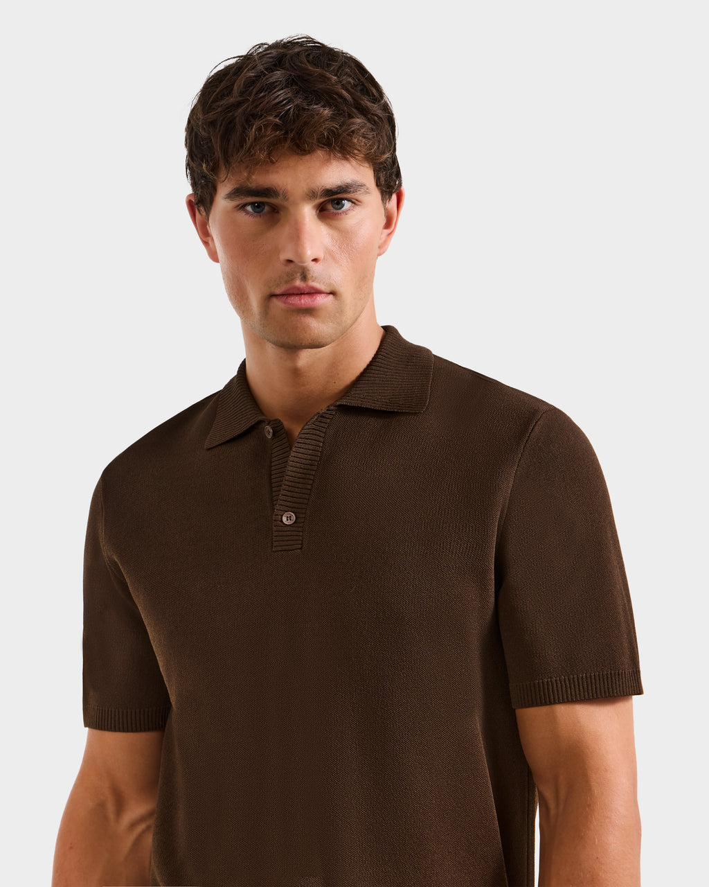 A young man with short, wavy brown hair wears the Rudsak MAISEN - 6524518 Mocha men's knit polo and stands against a plain light background, looking directly at the camera with a neutral expression.