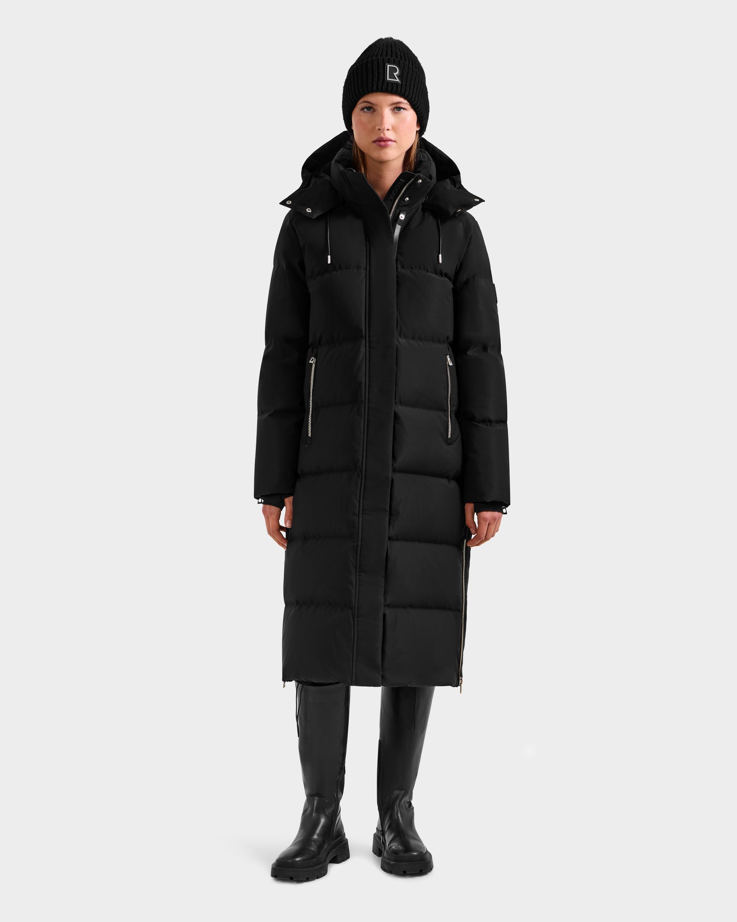 A person stands facing forward against a plain light background wearing the Rudsak MAO H - 8125603 Black women’s long down puffer with removable hood, paired with a black beanie, black boots, and black pants.