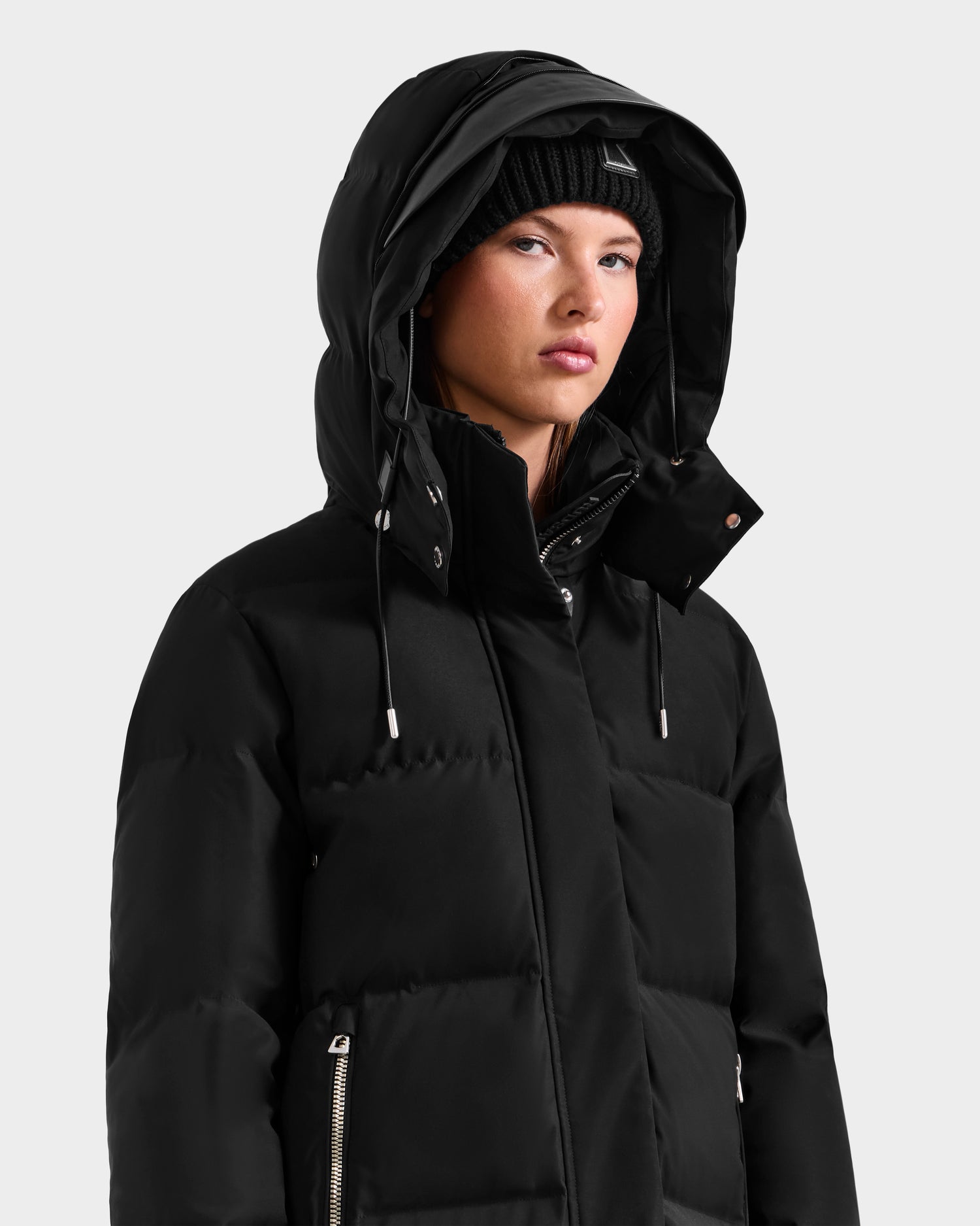A person wearing the Rudsak MAO H - 8125603 Black weather-resistant winter coat with a removable hood and a black knit beanie, looking slightly to the side against a plain light background.