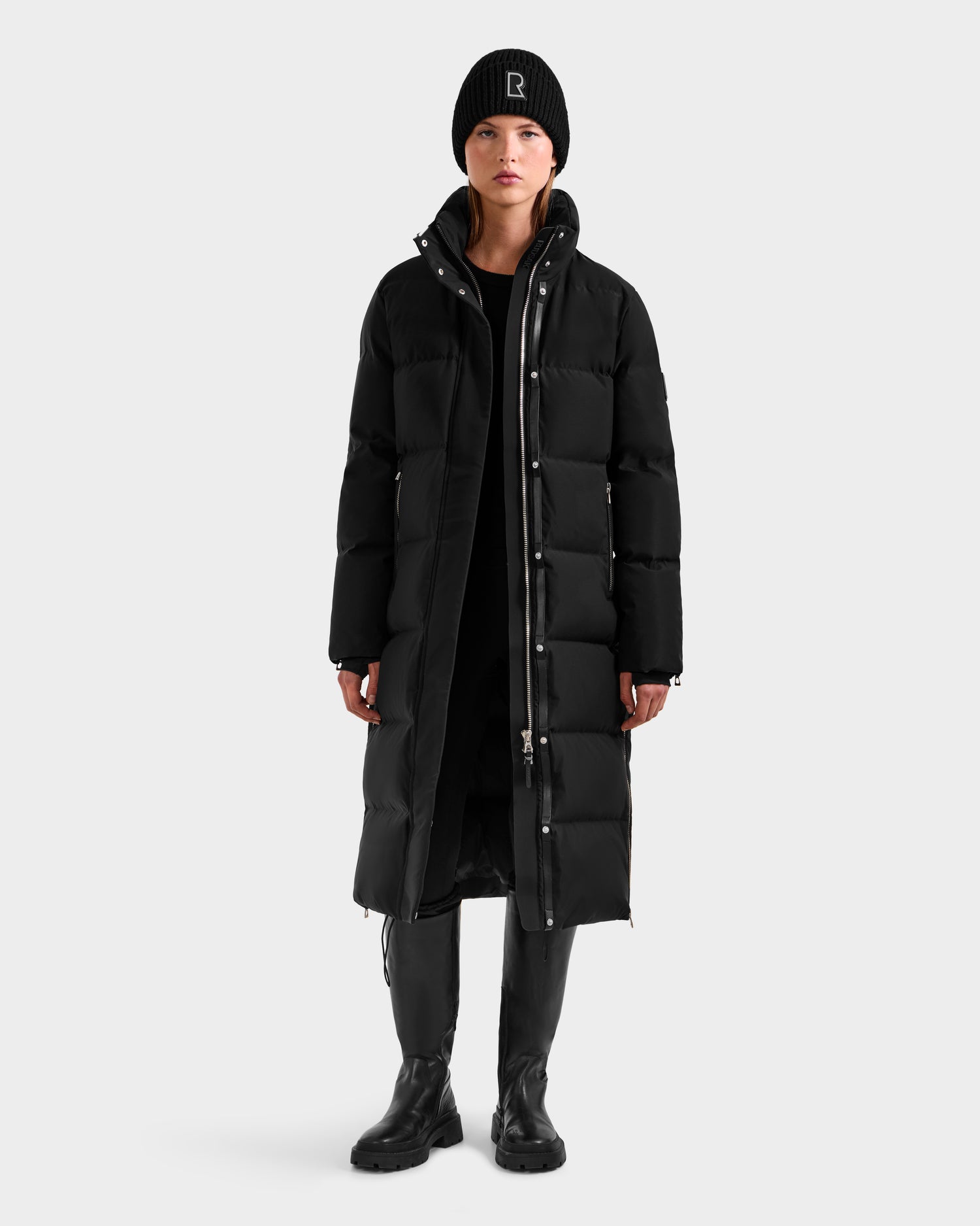 A person stands facing forward against a plain light background, wearing the Rudsak MAO H - 8125603 Black women’s long down puffer with a removable hood, paired with a black beanie, boots, and clothing.