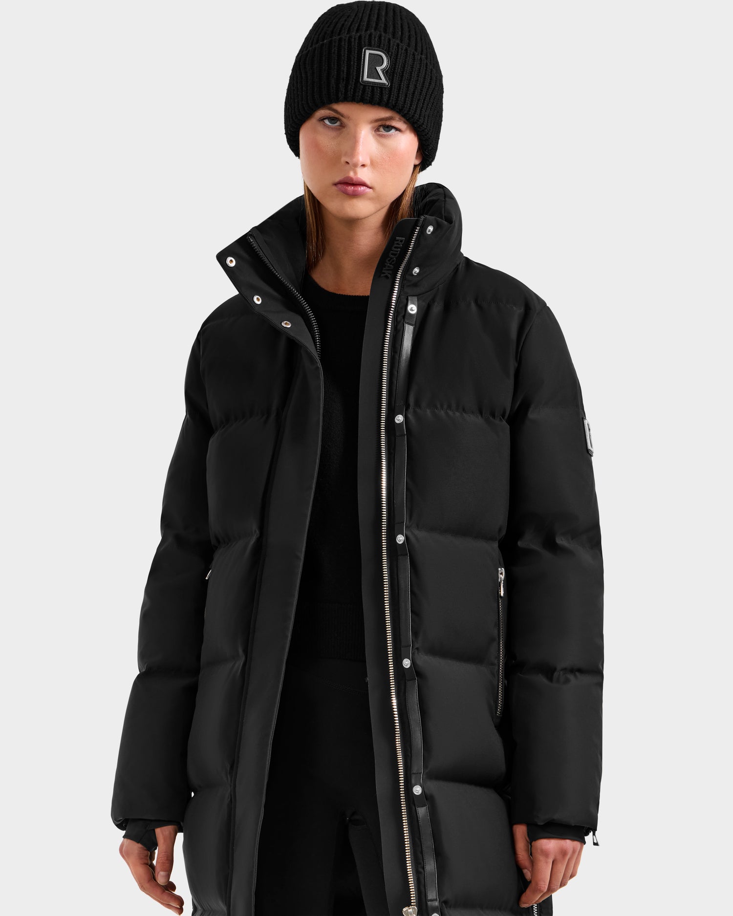 A woman with straight brown hair and a neutral expression stands against a plain light background, wearing the Rudsak MAO H - 8125603 Black women’s long down puffer with removable hood and a black knit beanie.