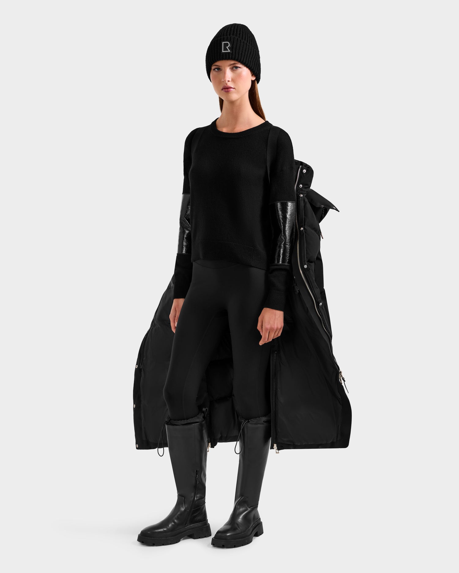 A woman faces forward in the Rudsak MAO H - 8125603 Black weather-resistant winter coat with a removable hood, paired with a black knit hat, sweater, leggings, and knee-high boots against a plain light background.