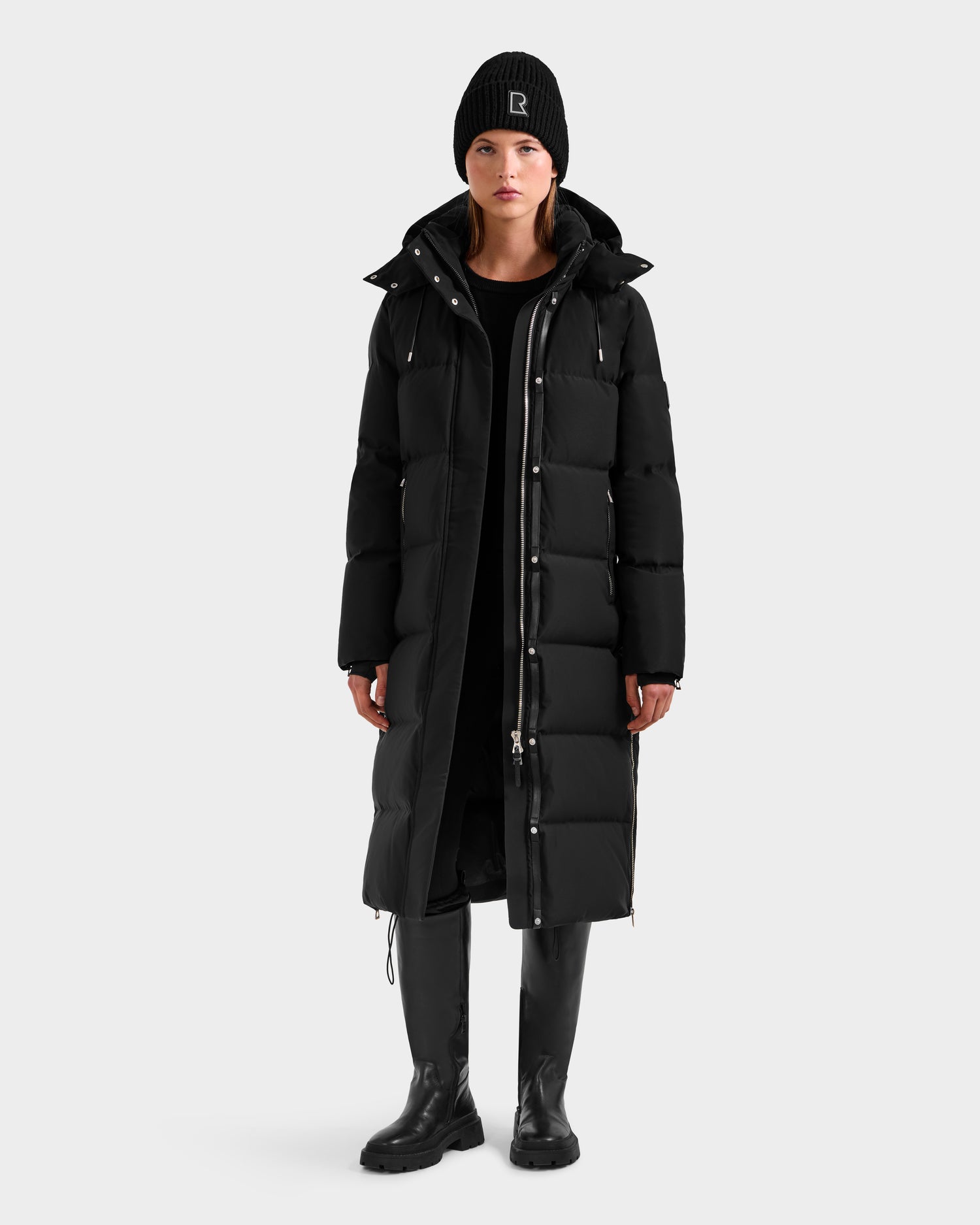 A person stands facing forward, wearing the Rudsak MAO H - 8125603 Black women’s long down puffer coat with removable hood, a black knit beanie, black dress, and tall black boots against a plain light gray background.