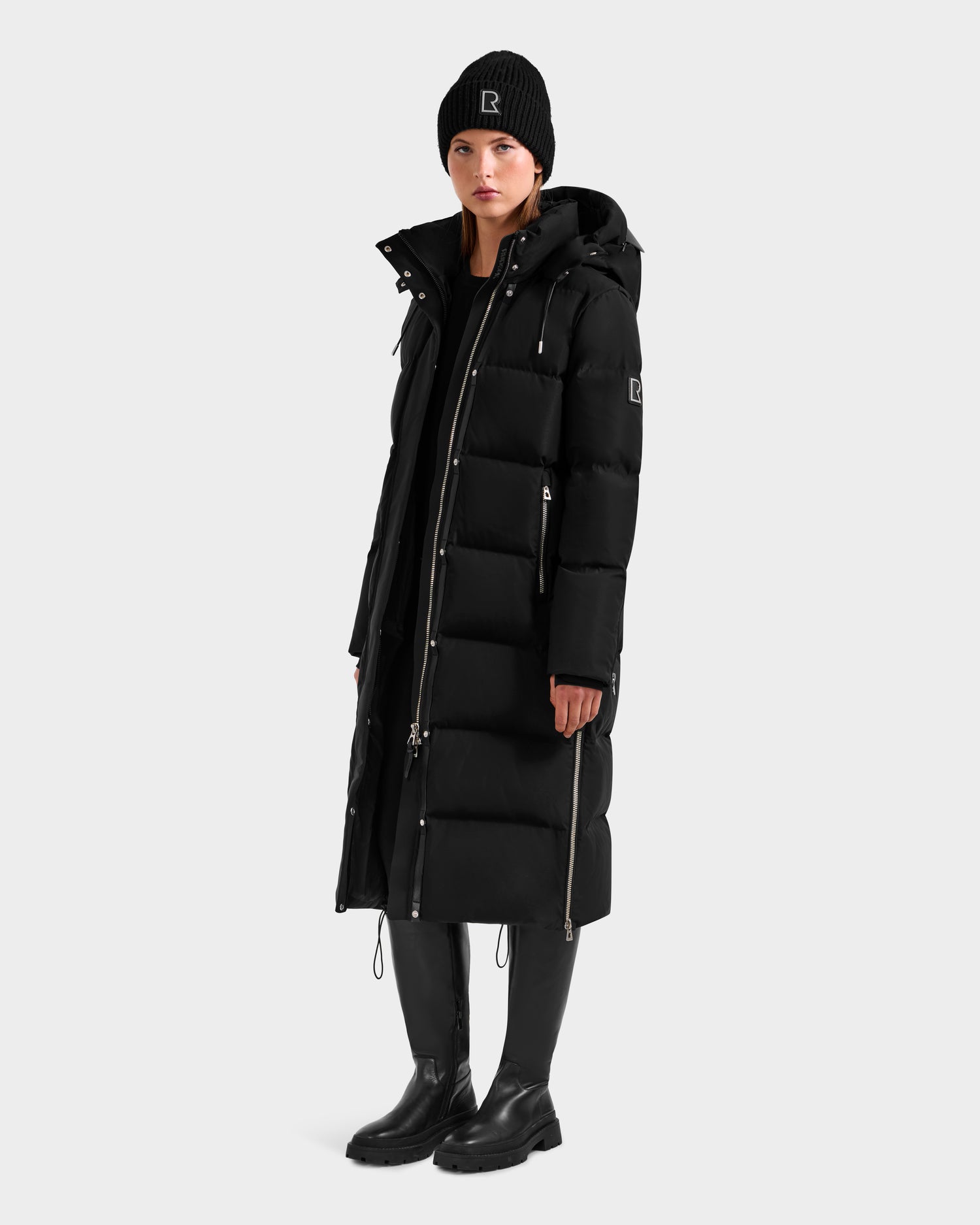 A woman stands against a light gray background wearing the Rudsak MAO H - 8125603 Black women’s long down puffer coat with removable hood, visible zippers, logo patch on the sleeve, black beanie, pants, and boots. The coat is weather-resistant.