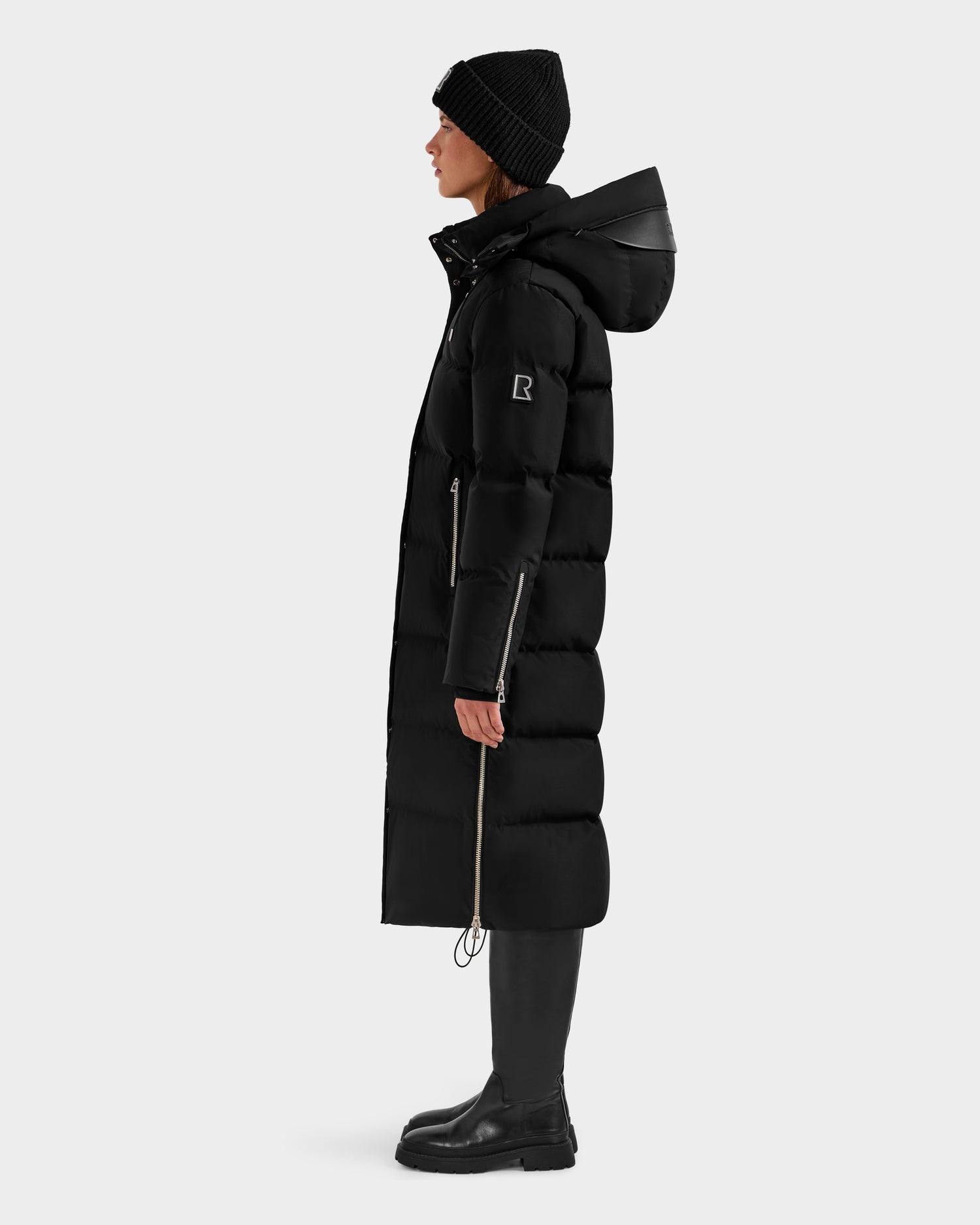 A person stands in profile against a white background wearing the Rudsak MAO H - 8125603 Black women’s long down puffer with removable hood, styled with a black beanie, black boots, and black pants.