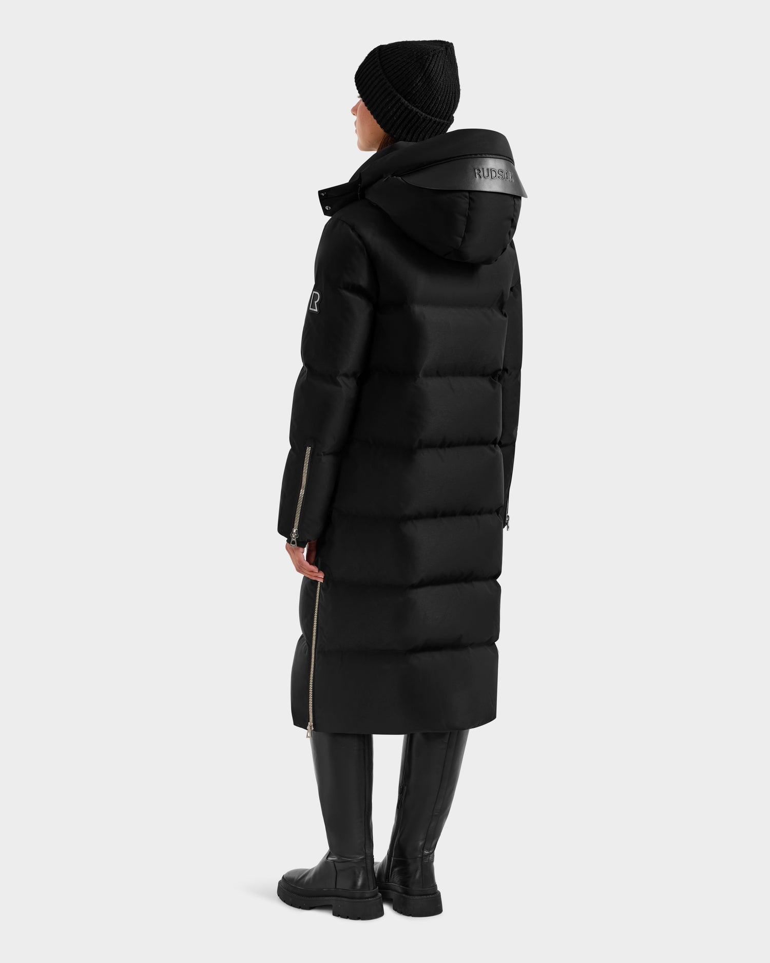 A person faces away, wearing the Rudsak MAO H - 8125603 Black women’s long down puffer with side zippers and a logo on the removable hood, paired with a black beanie, boots, and dark pants against a plain light gray background.