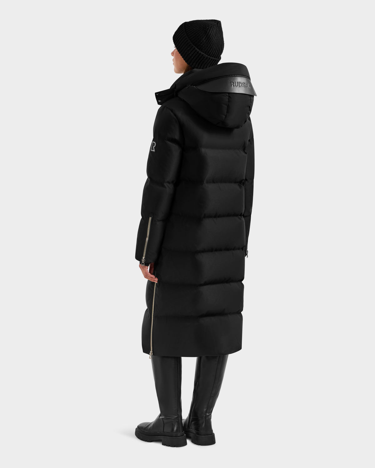 A person faces away, wearing the Rudsak MAO H - 8125603 Black women’s long down puffer with side zippers and a logo on the removable hood, paired with a black beanie, boots, and dark pants against a plain light gray background.