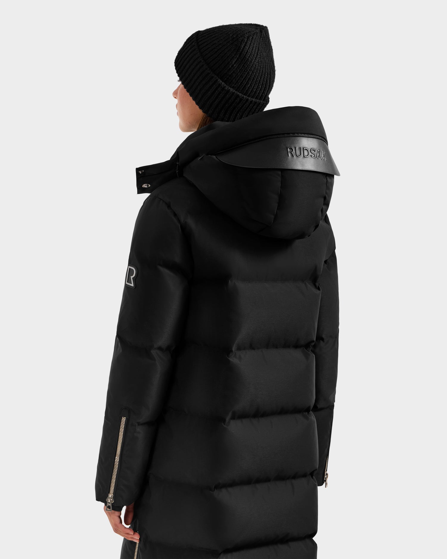 A person wearing the Rudsak MAO H - 8125603 Black, a weather-resistant winter coat with a hood featuring the RUDSAK logo, plus a black knit beanie. Seen from the back, the coat has a sleeve zipper and delivers reliable warmth for winter days.