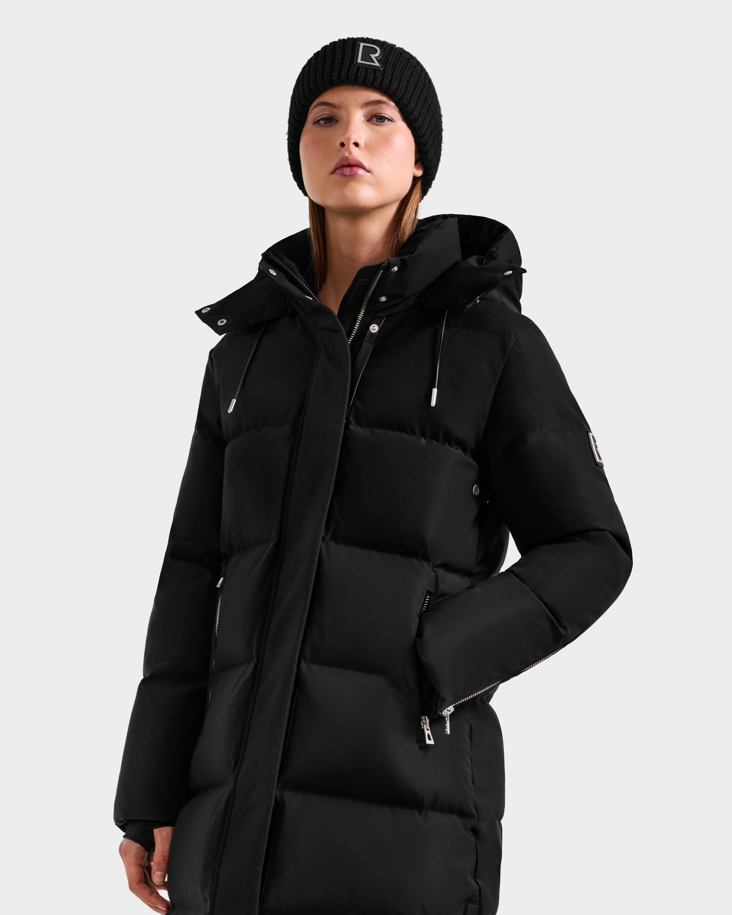 A woman wears the Rudsak MAO H - 8125603 Black weather-resistant winter coat with removable hood and a black knit beanie, standing confidently with one hand in her pocket against a plain background.