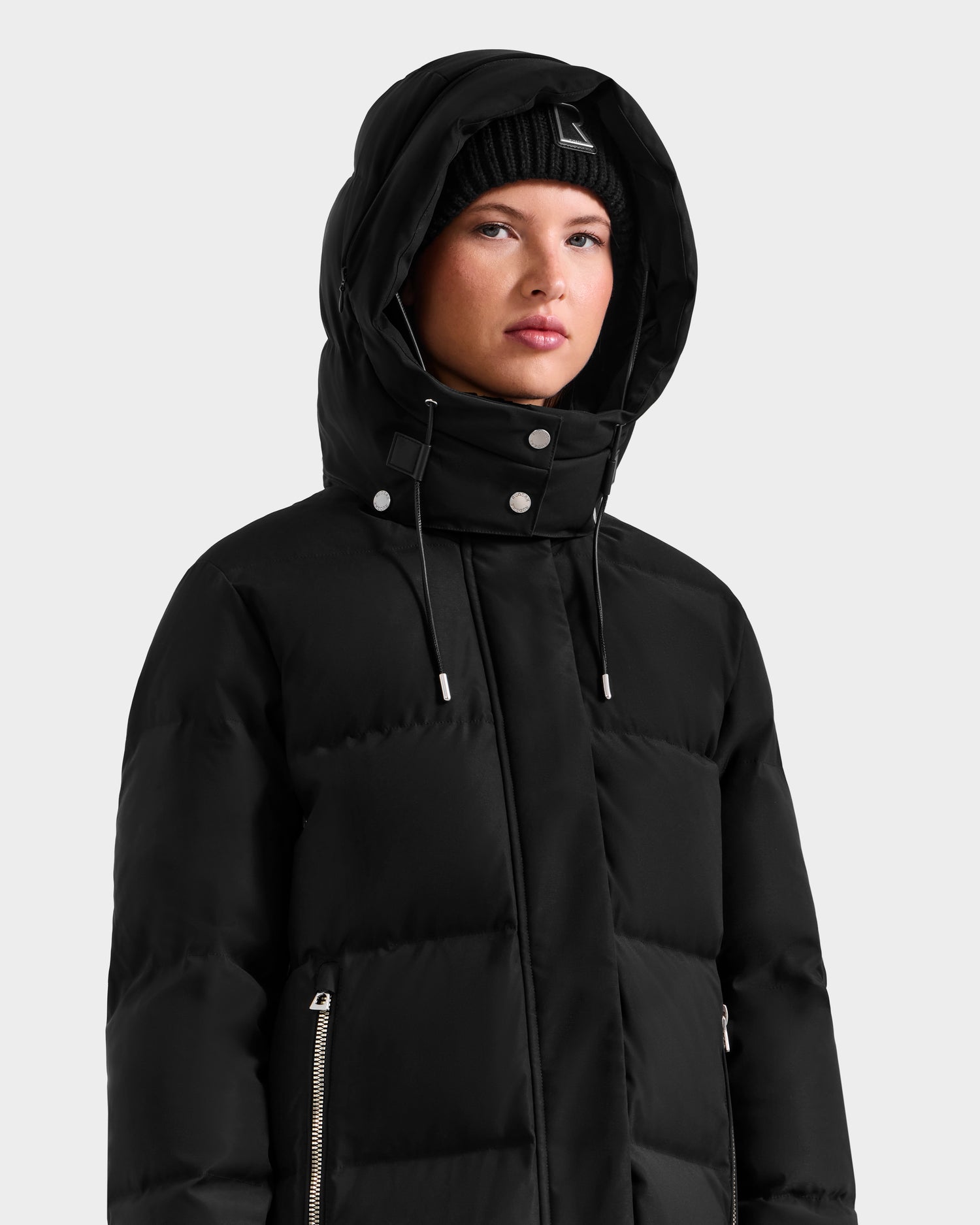 A woman wearing the Rudsak MAO H - 8125603 Black women’s long down puffer with removable hood and a black knit beanie faces forward against a plain light background.