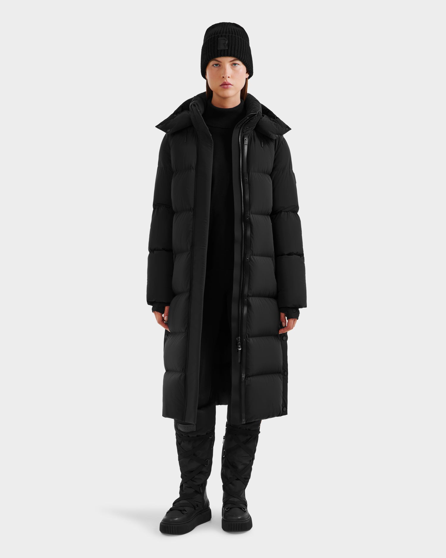A person stands facing forward wearing the Rudsak MAO - 8124780 Black women’s long down coat, paired with a black beanie, pants, and quilted boots—perfect winter wear—against a light gray background. The coat is wind and water resistant.