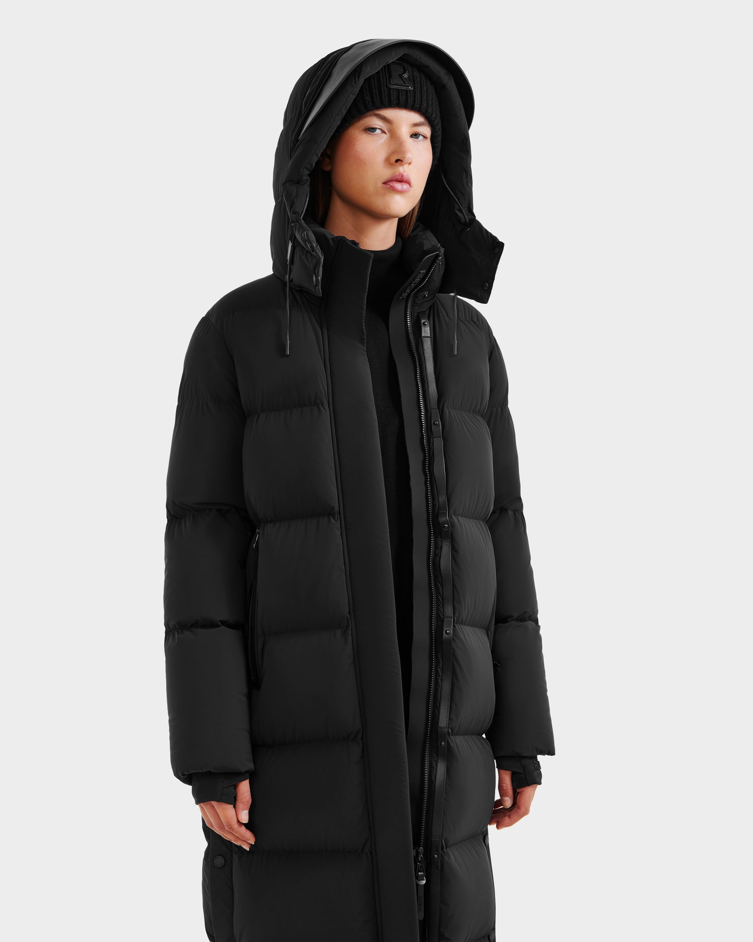 A person confidently embraces winter style, wearing the Rudsak MAO - 8124780 Black women’s long down coat with a hood, paired with a black beanie and top, against a plain light background.