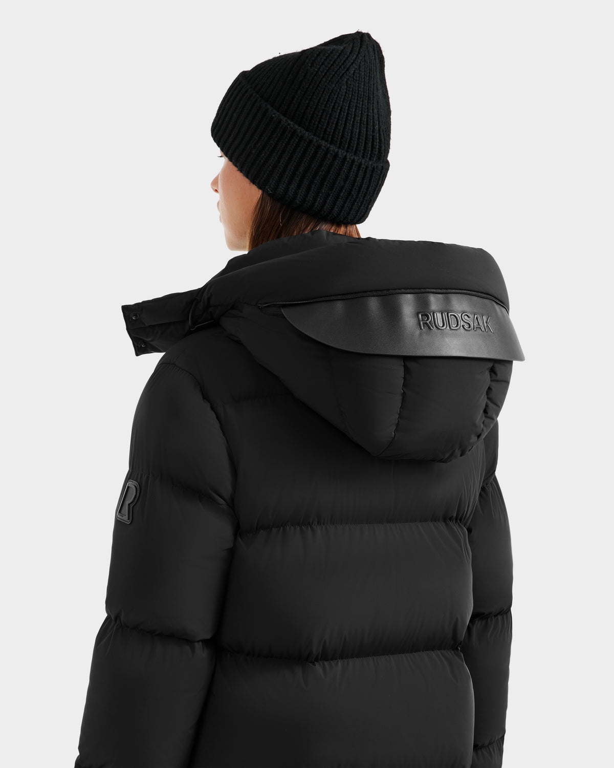 A person wearing the Rudsak MAO - 8124780 Black knit beanie and womens long down coat with RUDSAK embossed on the hood, shown from behind against a light background—an ideal look for stylish winter wear.