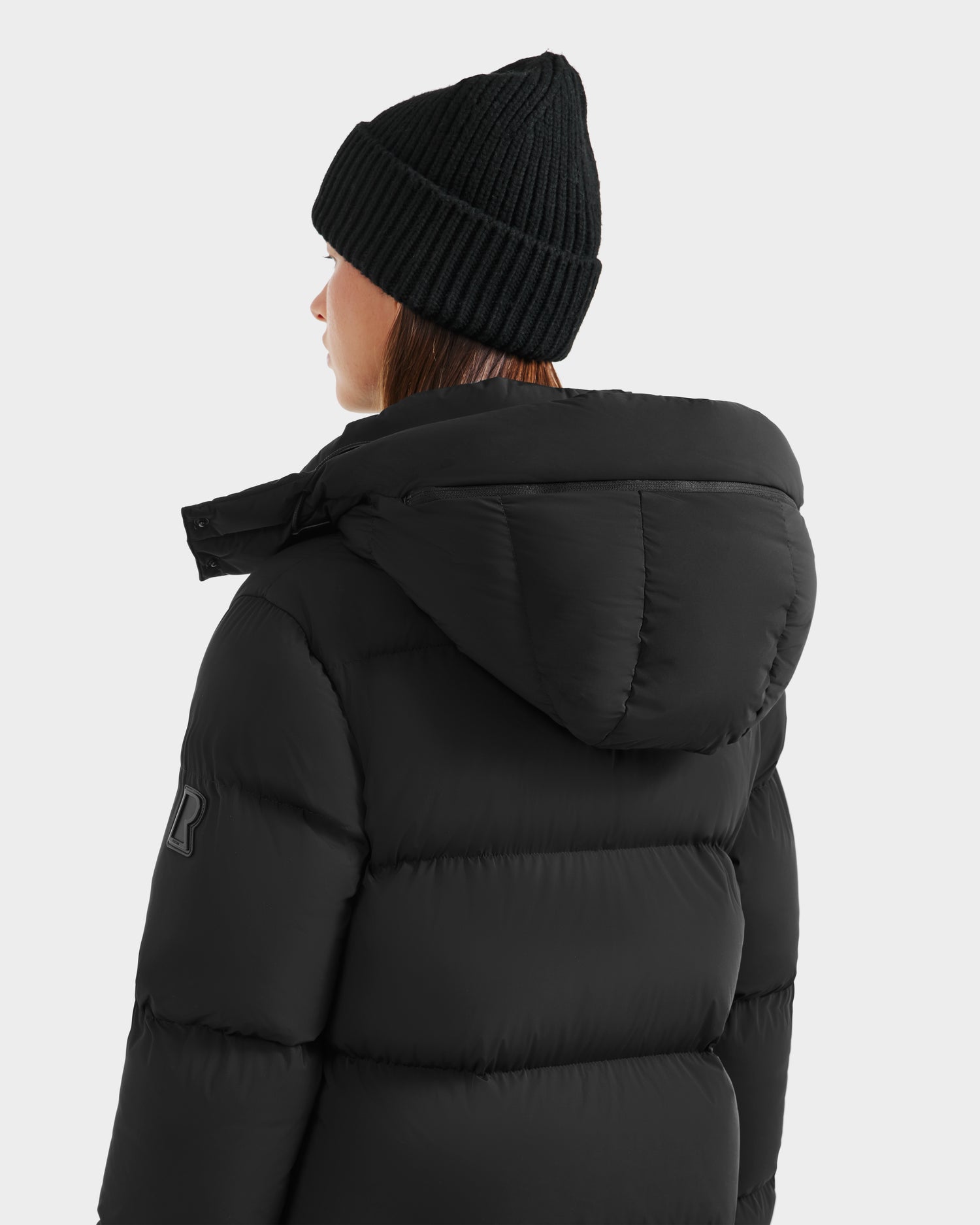 A person wearing a Rudsak MAO - 8124780 Black puffer jacket with a hood and a black knit beanie, seen from the back and slightly to the side, against a plain light gray background.