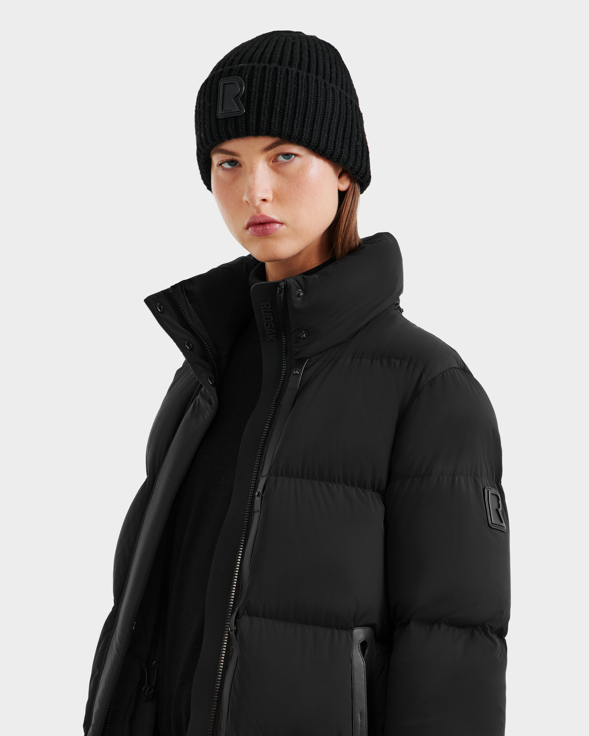 A person in the Rudsak MAO - 8124780 Black wind and water-resistant puffer jacket with a high collar and a black beanie stands against a light gray background, looking directly at the camera with a neutral expression.